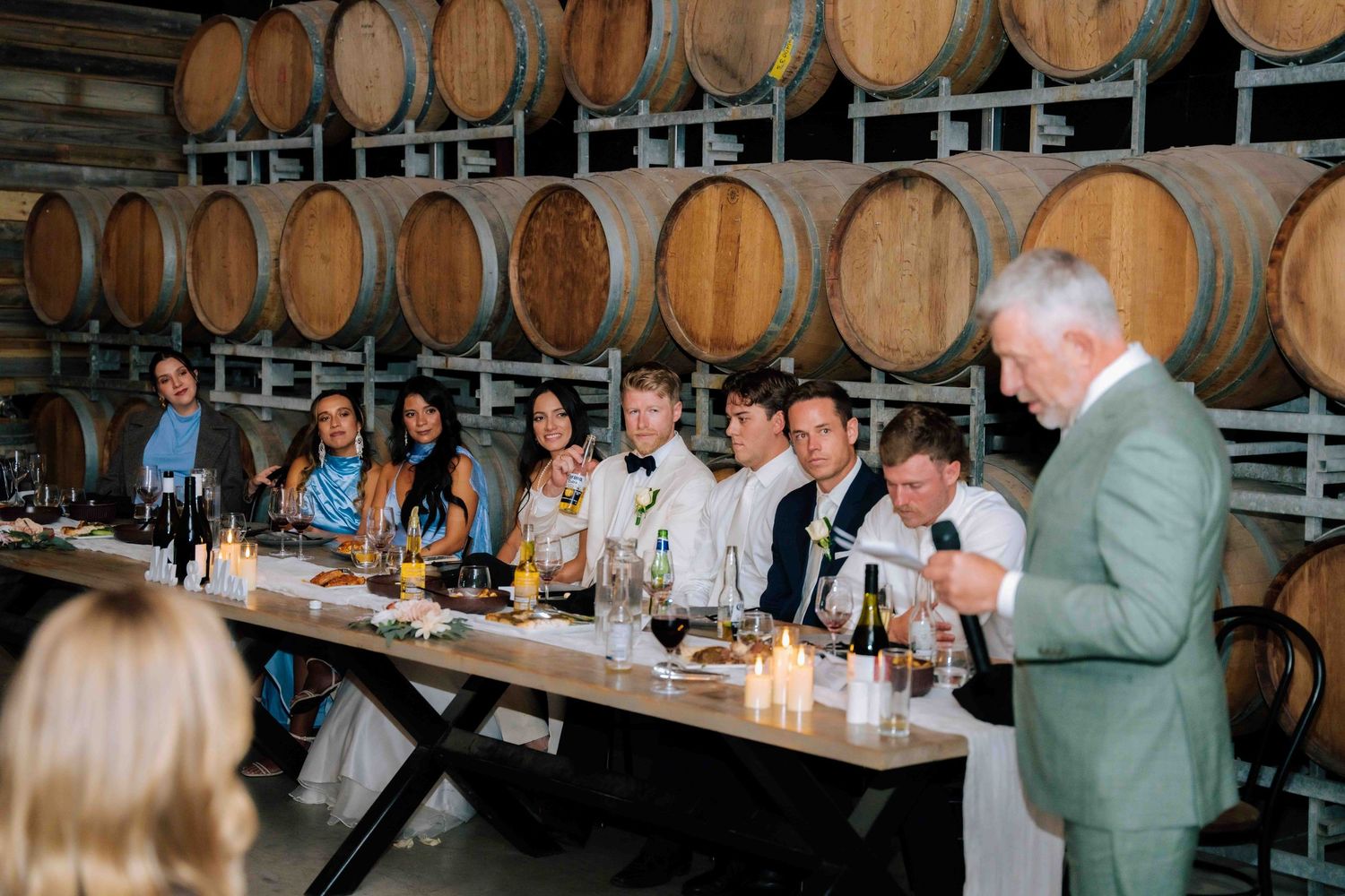 Wedding guests seated at long table surrounded by wine barrels during reception dinner speech.