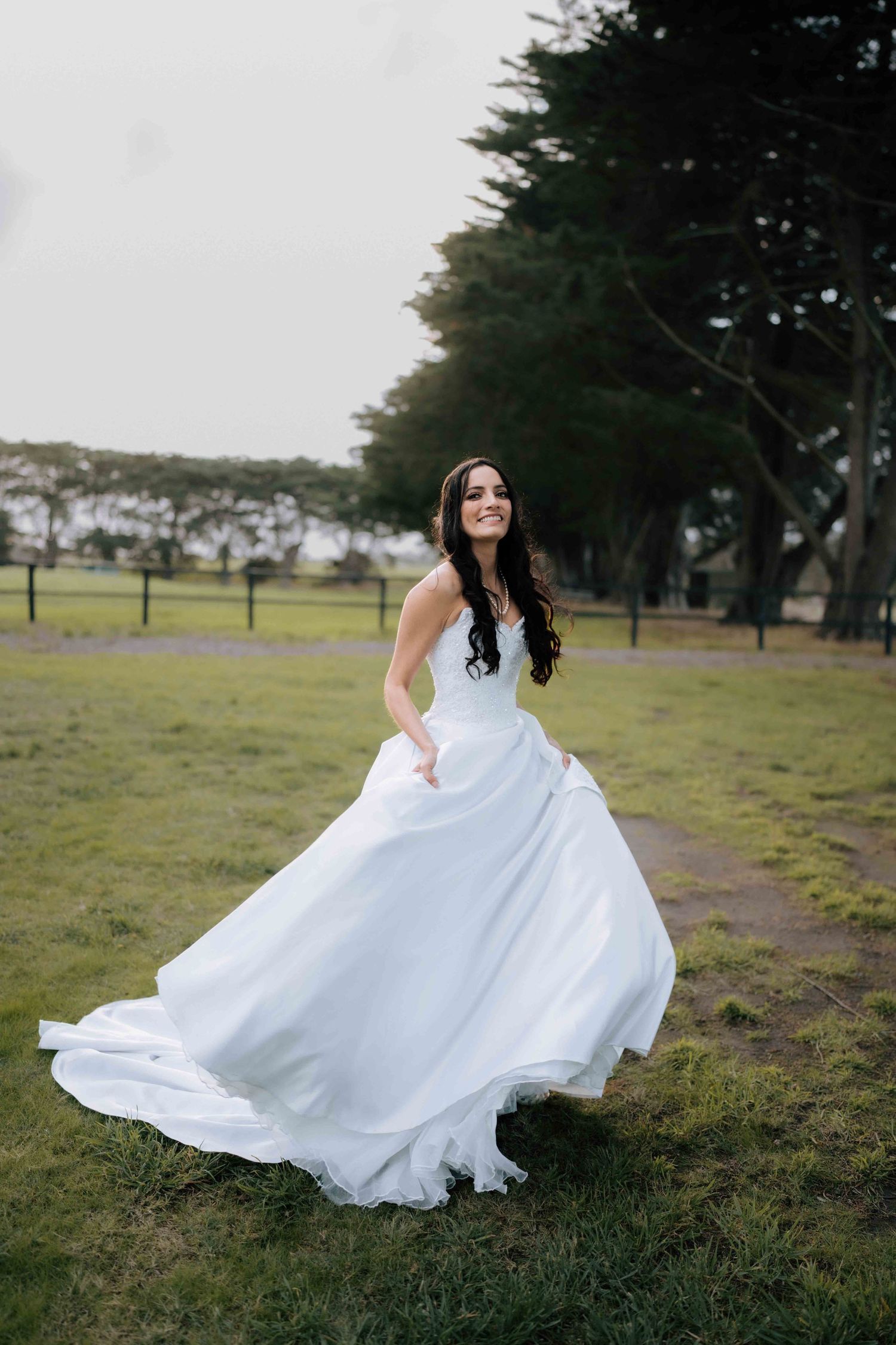 Bride in a flowing white ball gown twirling outdoors on green grass with trees in the background.