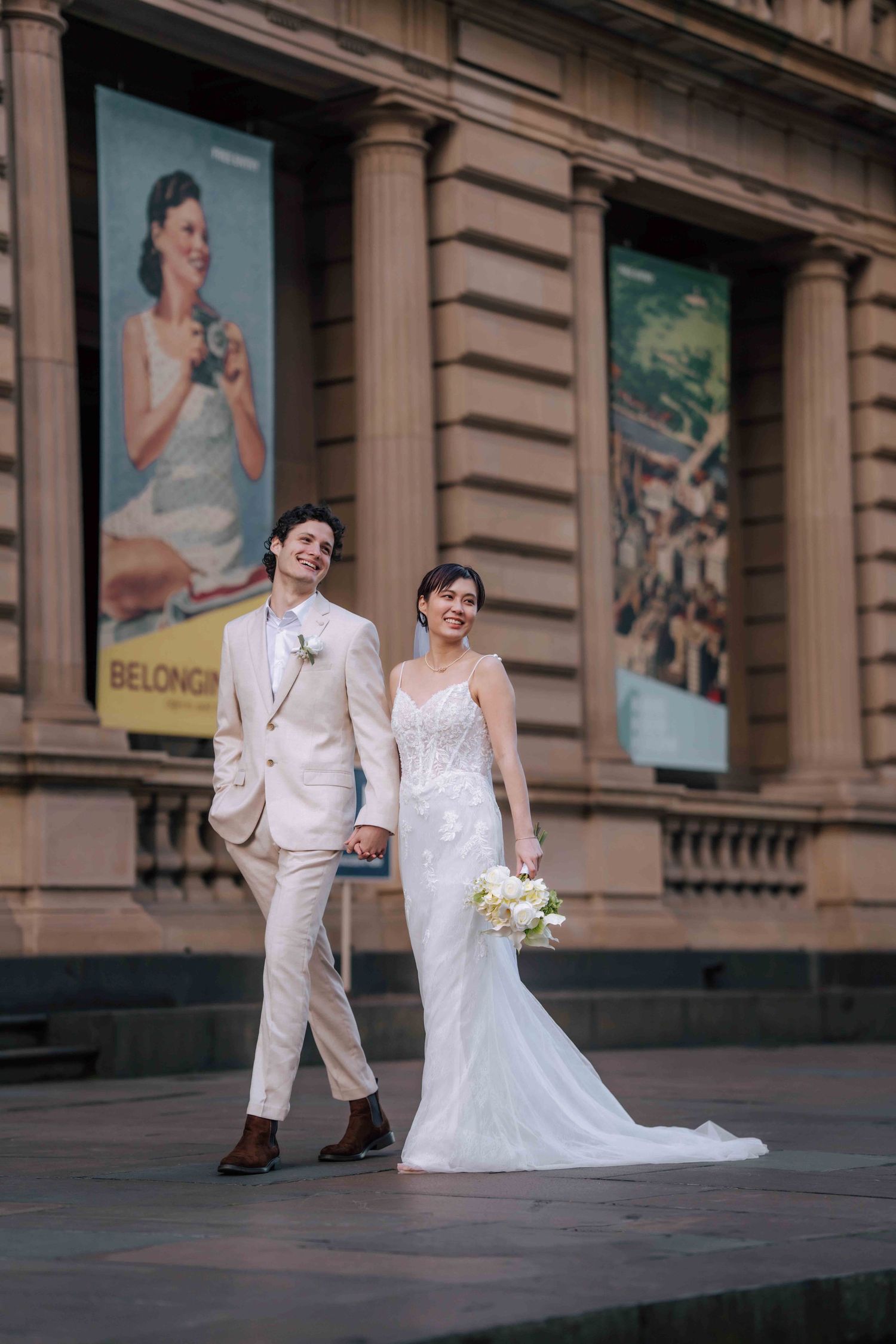 Smiling bride and groom walk hand in hand outside heritage building adorned with vintage travel posters.