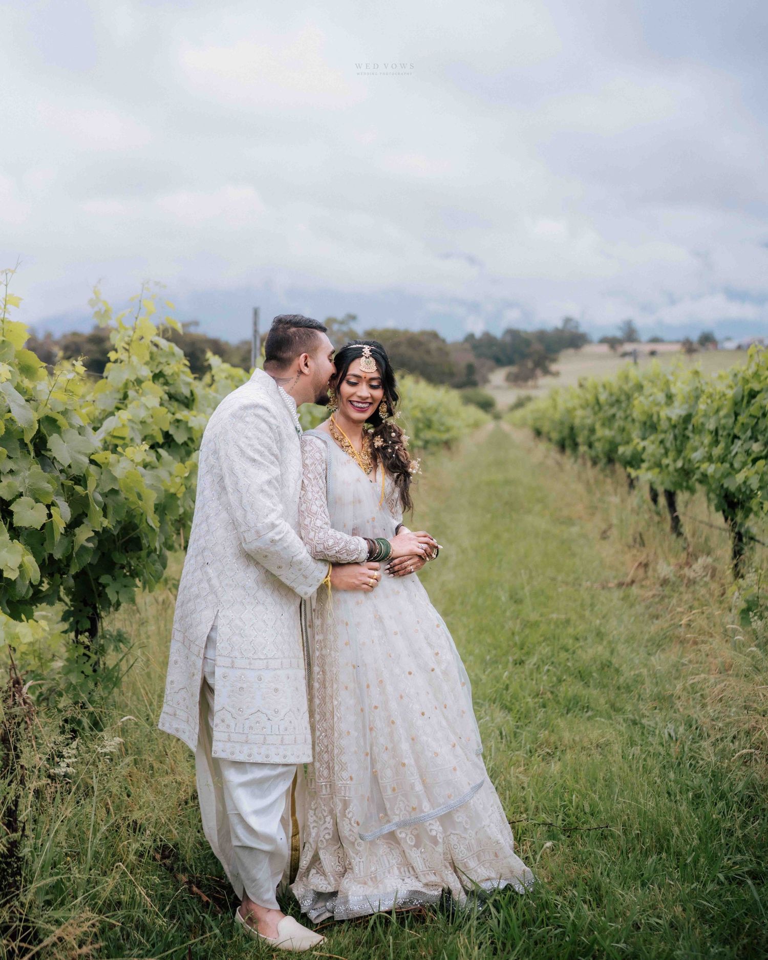 Smiling Indian bride and groom in white outfits share a romantic moment in vineyard rows.