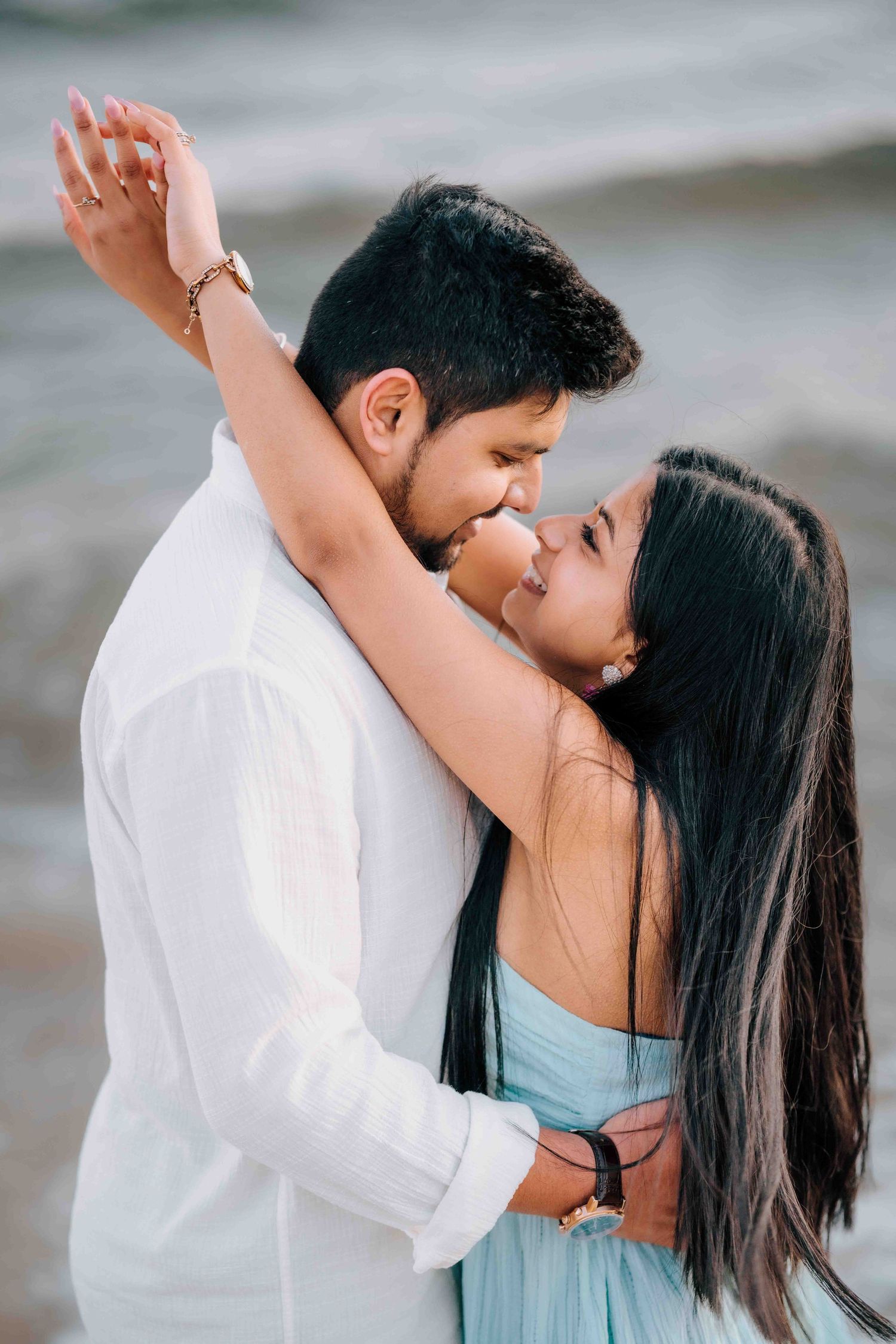 Romantic couple almost kissing on beach, woman in light blue dress with arm raised, ocean backdrop.