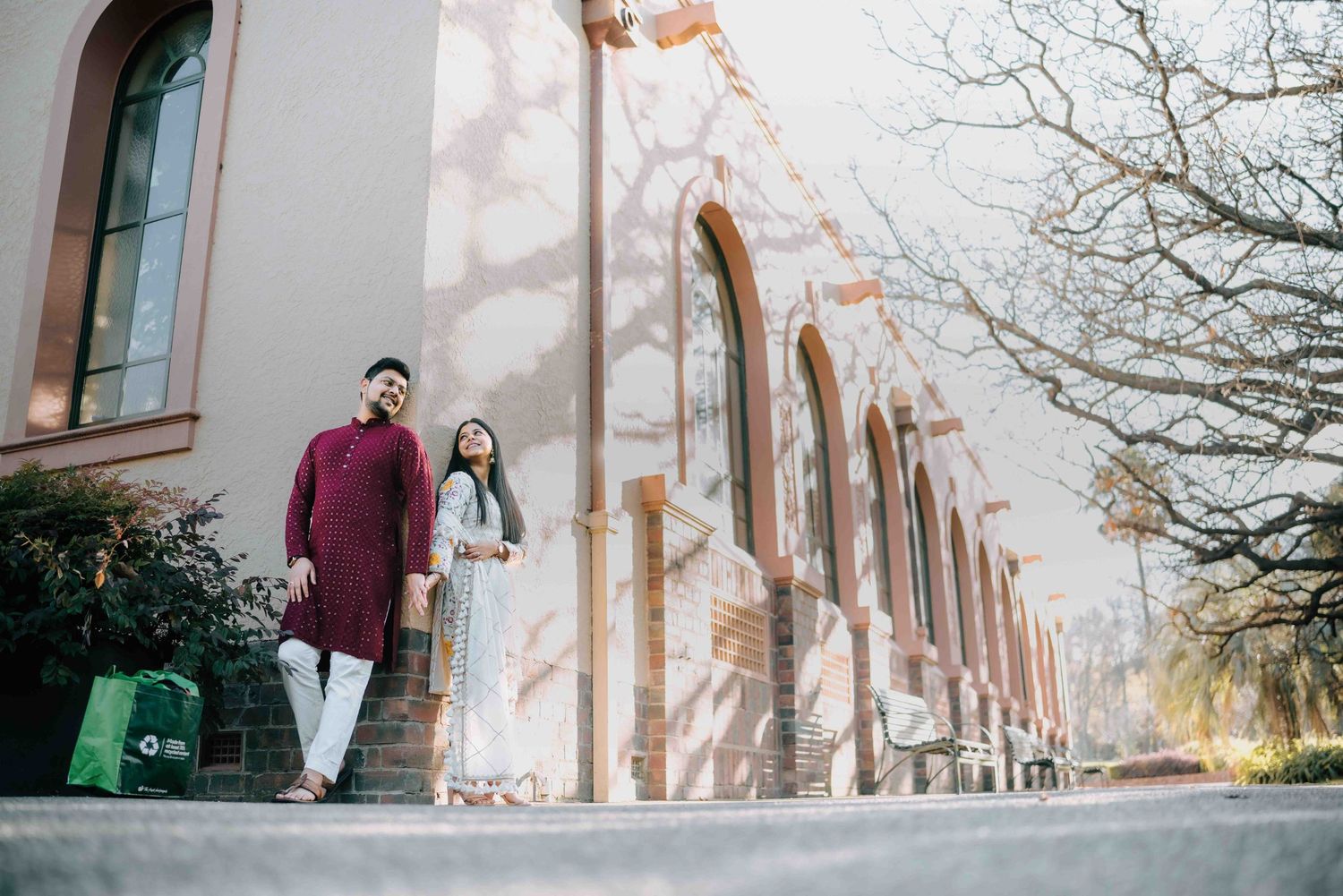 Couple in traditional South Asian attire walking beside sunlit historic red brick arched building.