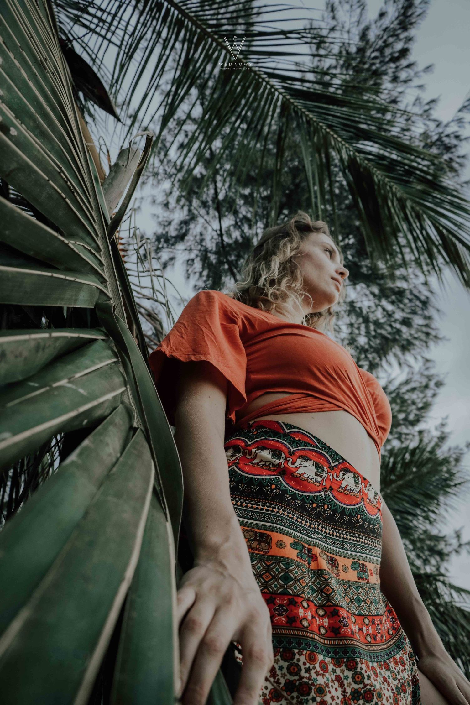 Low angle view of woman in orange crop top standing among lush tropical palm trees.