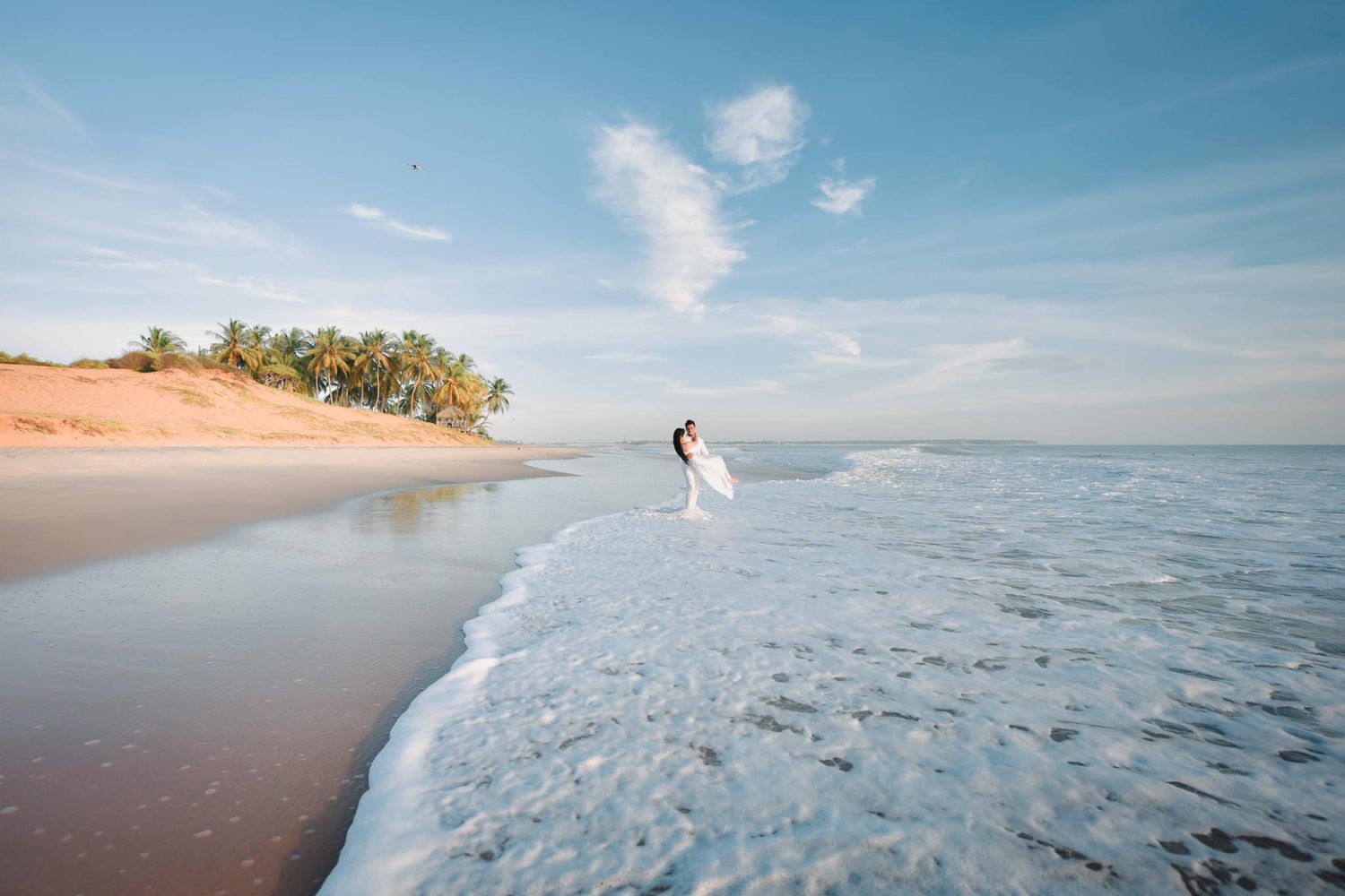 Bride and groom walking along a stunning tropical beach shoreline with palm trees and blue sky.