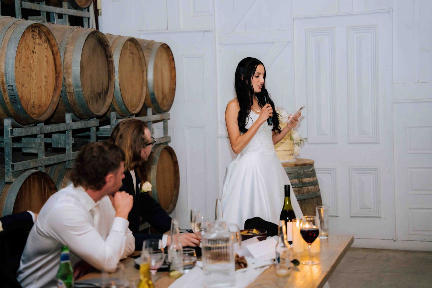 Bride in white gown gives a heartfelt speech holding a microphone at a candlelit winery wedding reception.