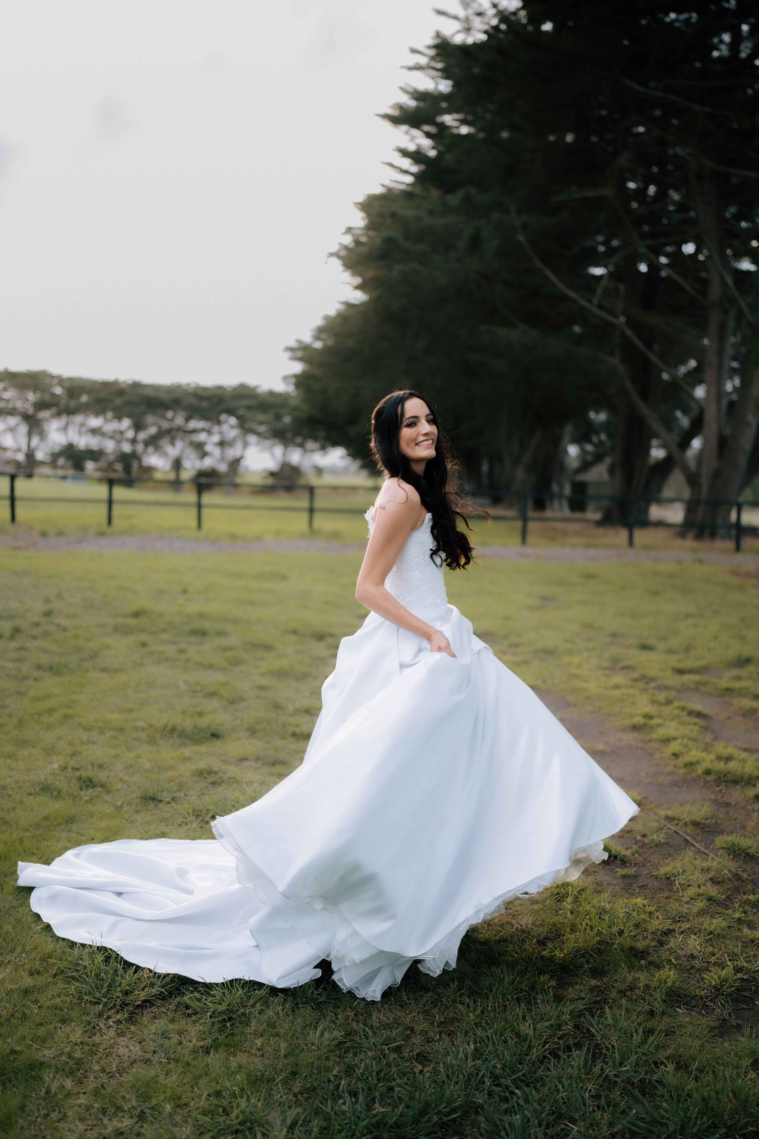 Smiling bride in a flowing white ball gown twirling outdoors on green grass with trees in the background.