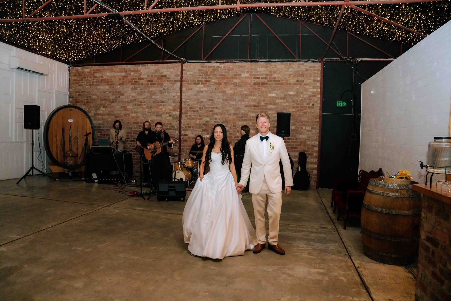 Bride and groom entering wedding reception in rustic barn with fairy lights and wine barrels.