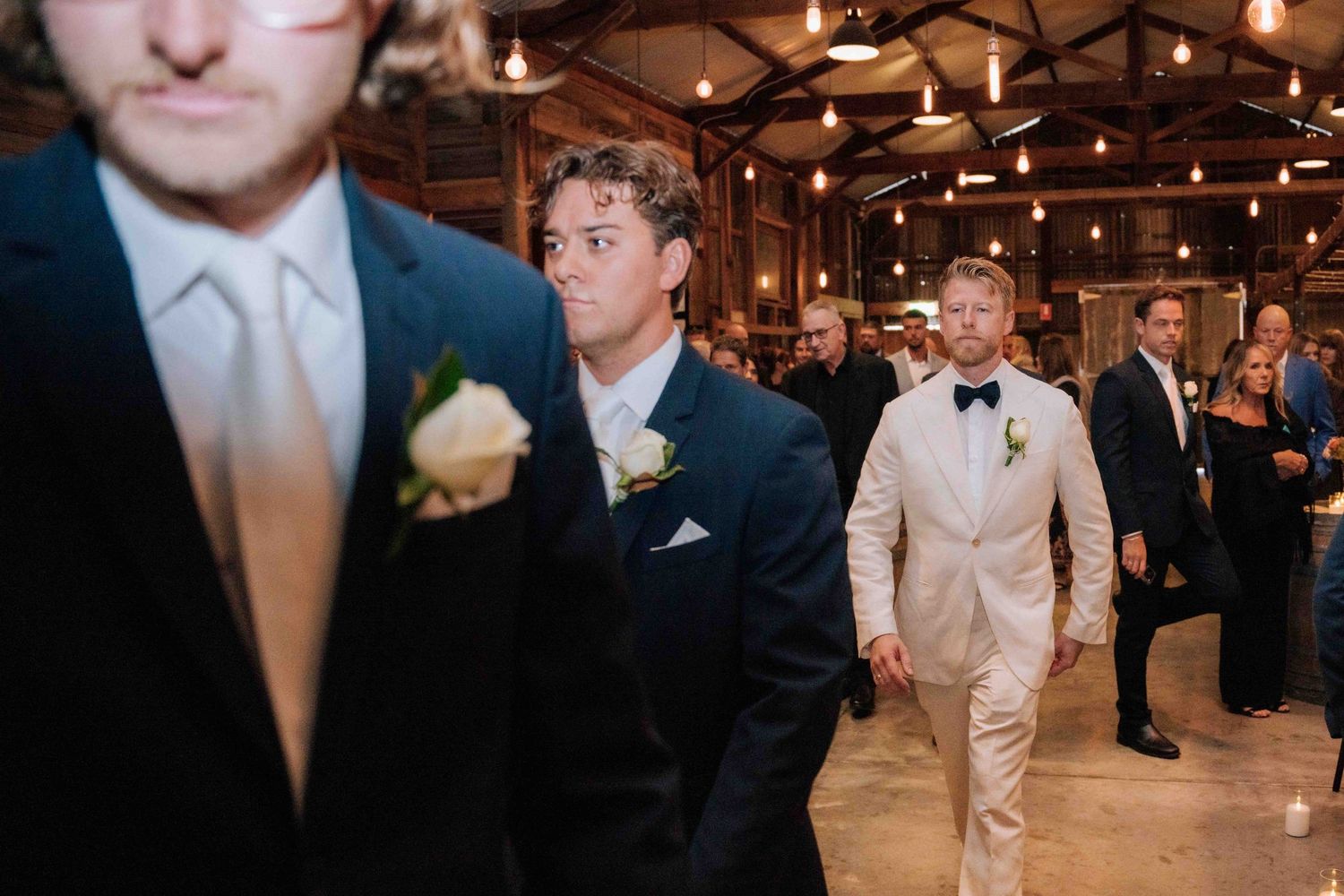Groom in cream tuxedo walks down rustic barn aisle with groomsmen in navy suits during wedding ceremony.