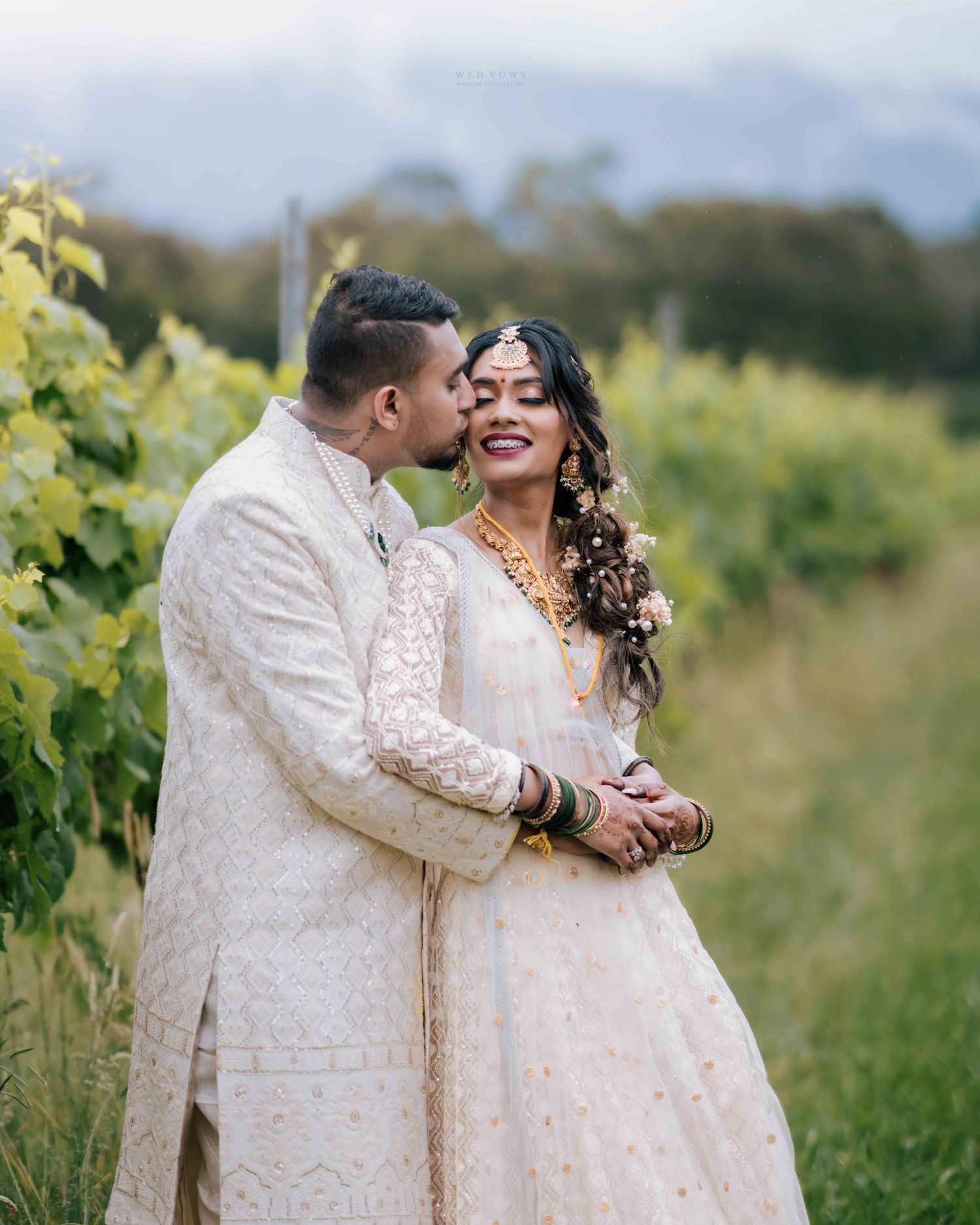 Indian groom in white sherwani kissing bride's cheek while she smiles in lush green vineyard.