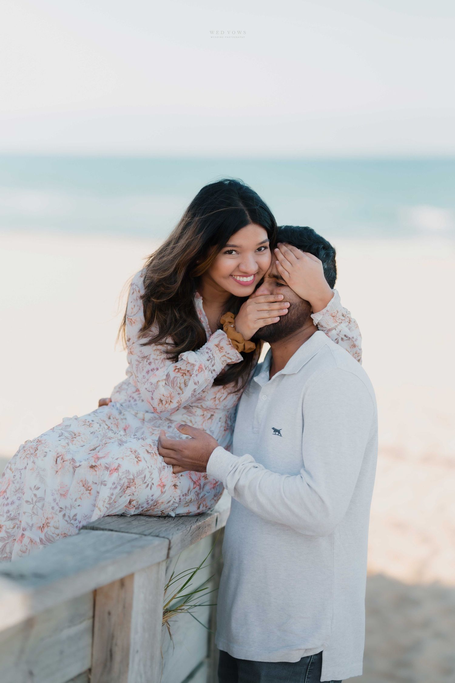 Smiling woman playfully covering her partners eyes while sitting on a beach boardwalk railing.