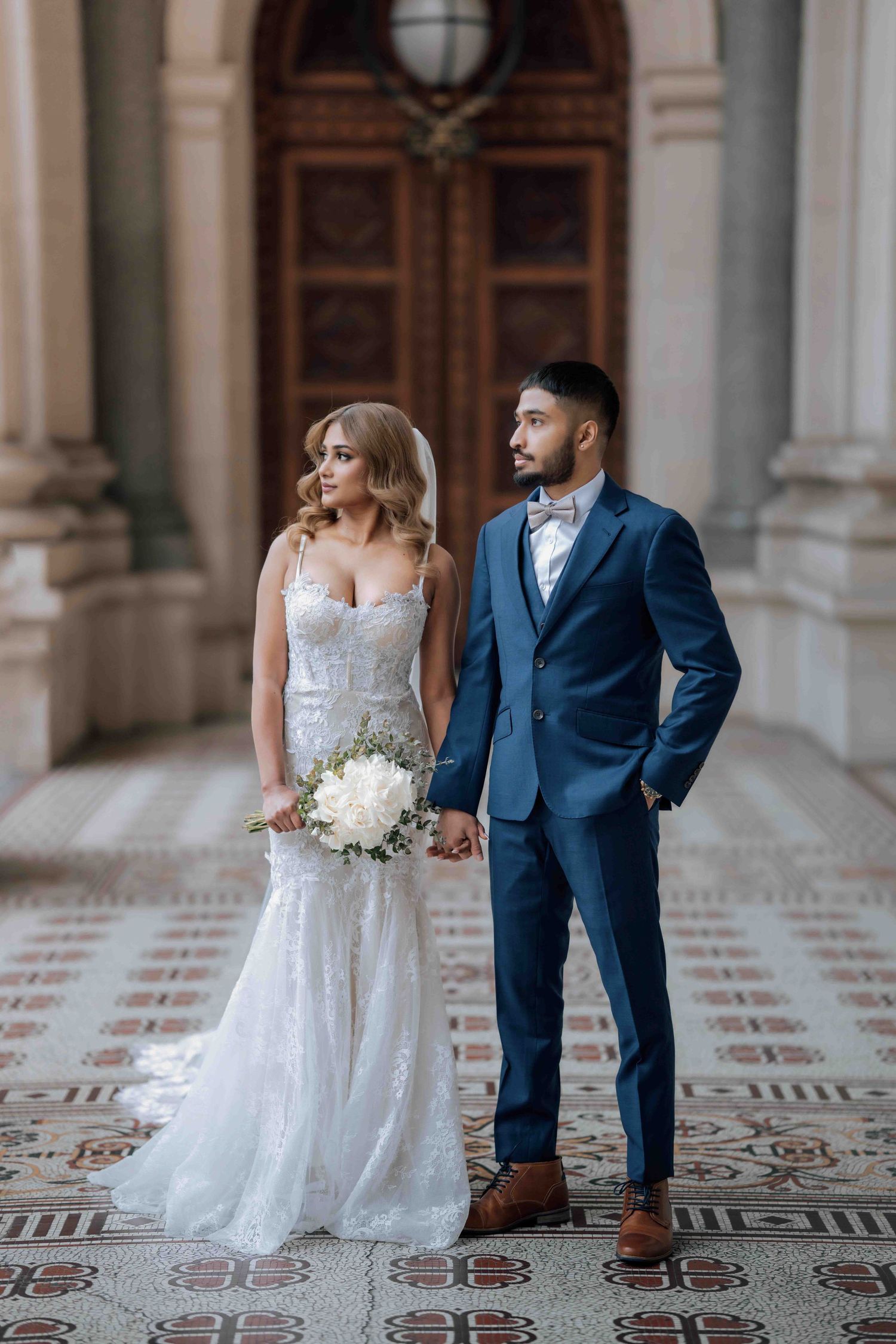 Bride holding white bouquet and groom in navy suit holding hands inside ornate building.