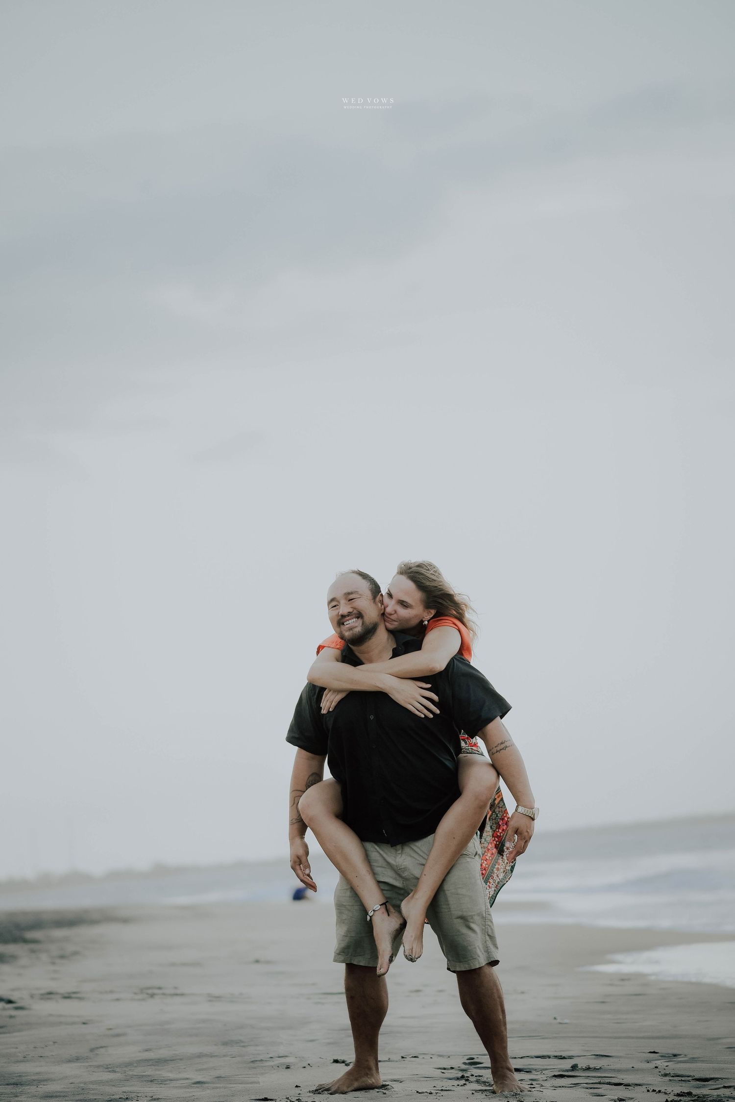 Man giving woman a piggyback ride on a sandy beach, both laughing with ocean in background.