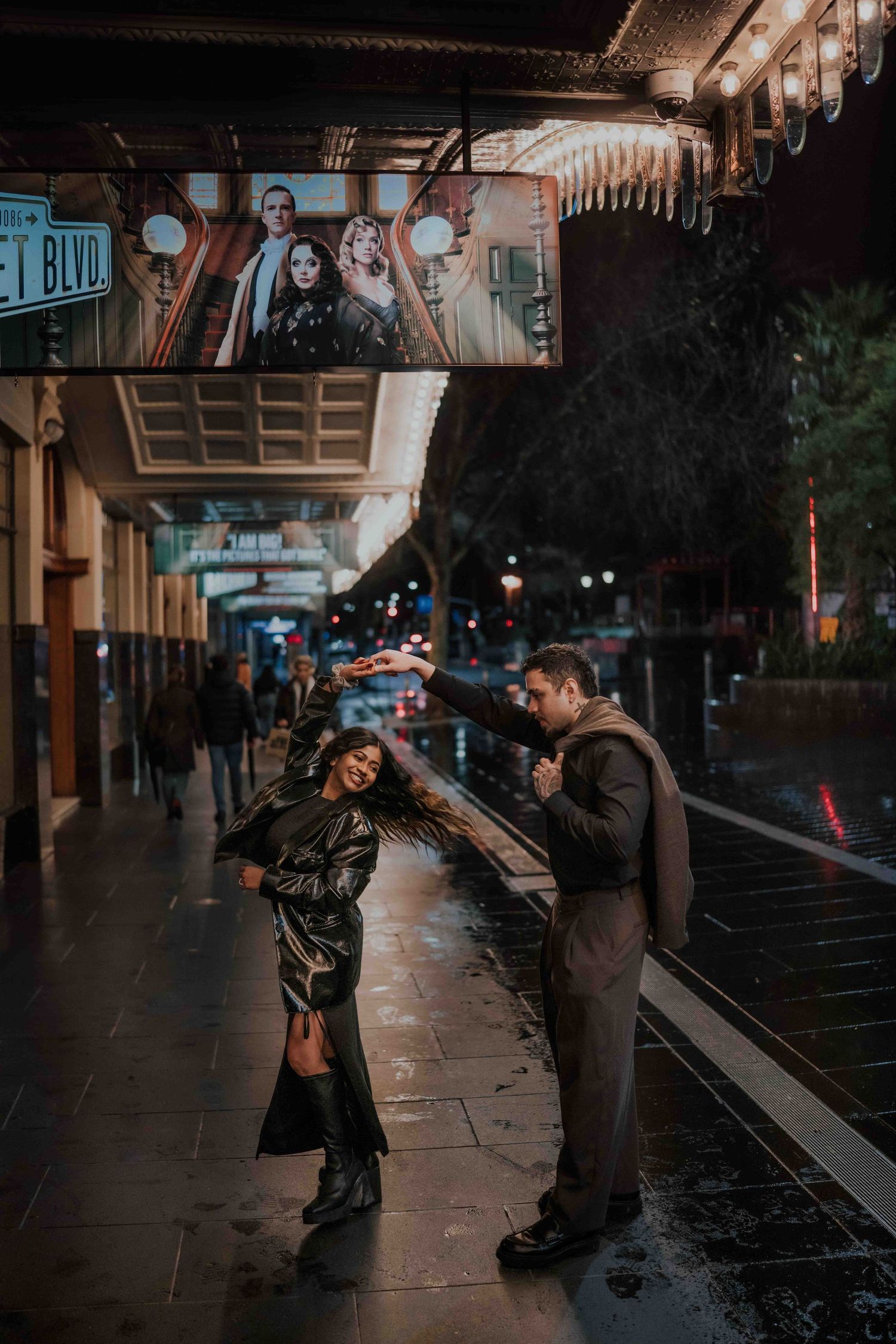 Woman twirling in the rain on a city street at night while her partner watches adoringly.