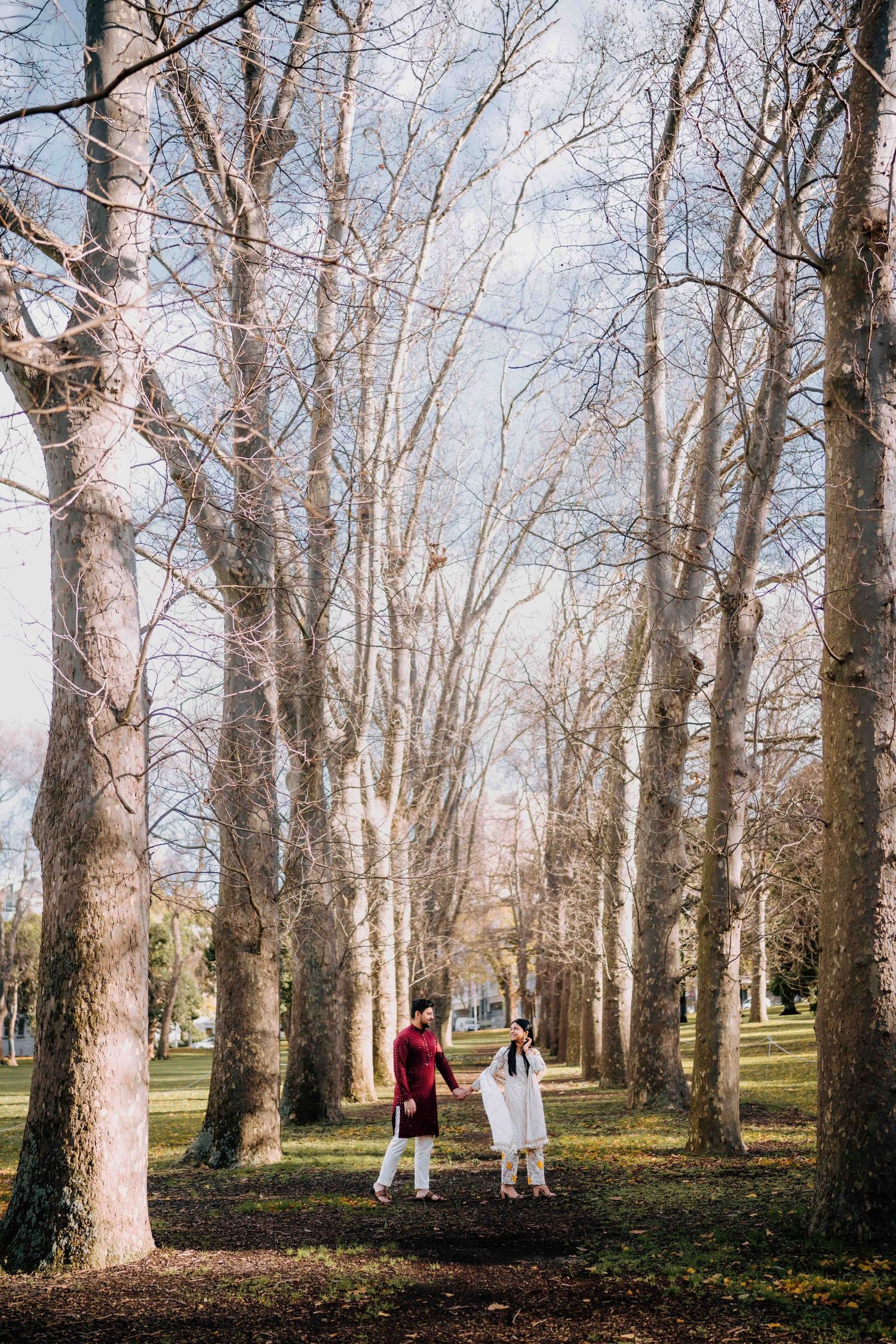 South Asian couple in traditional dress holding hands among tall bare trees in scenic park.