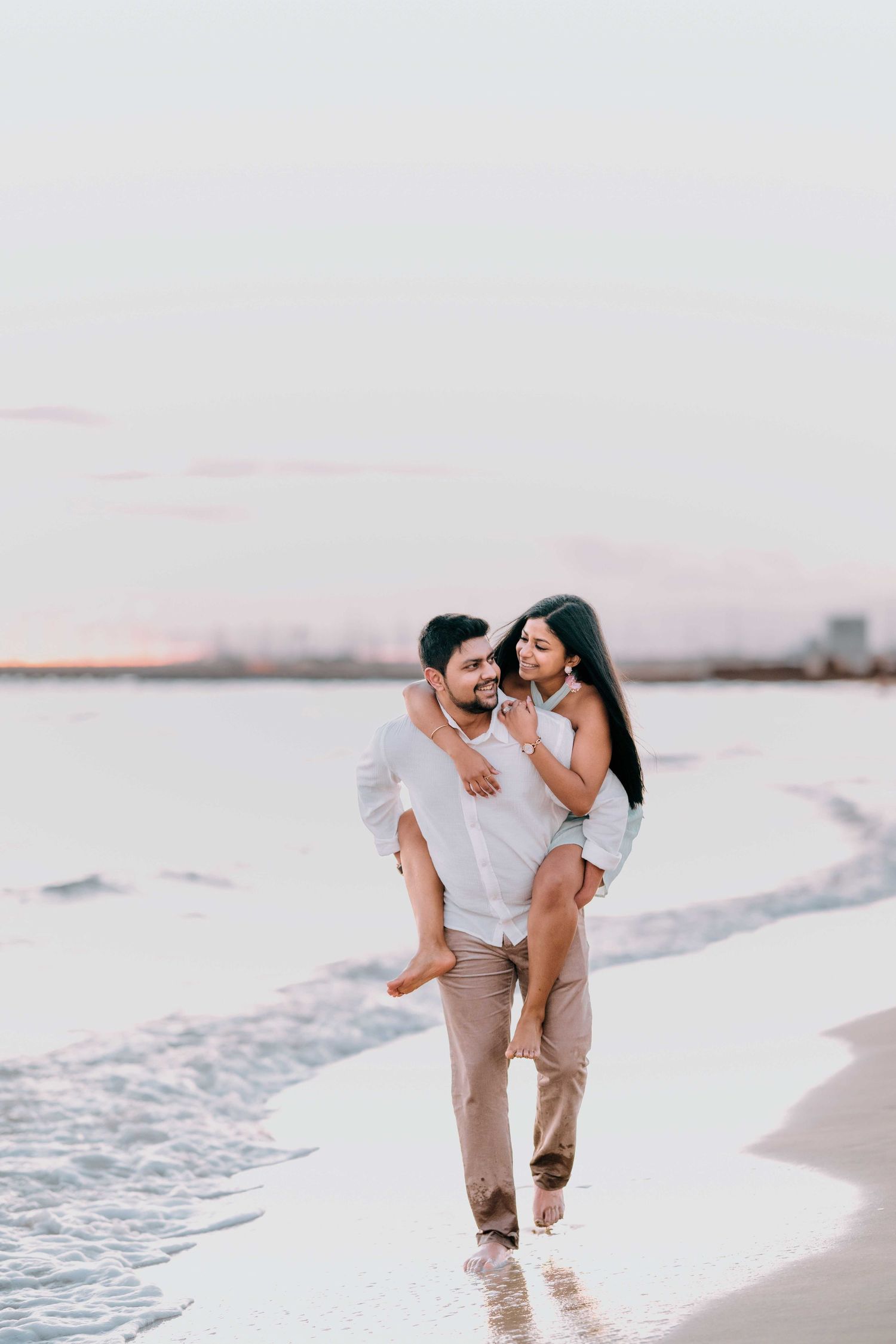 Happy couple at beach sunset, man carrying woman piggyback style while walking through ocean waves.