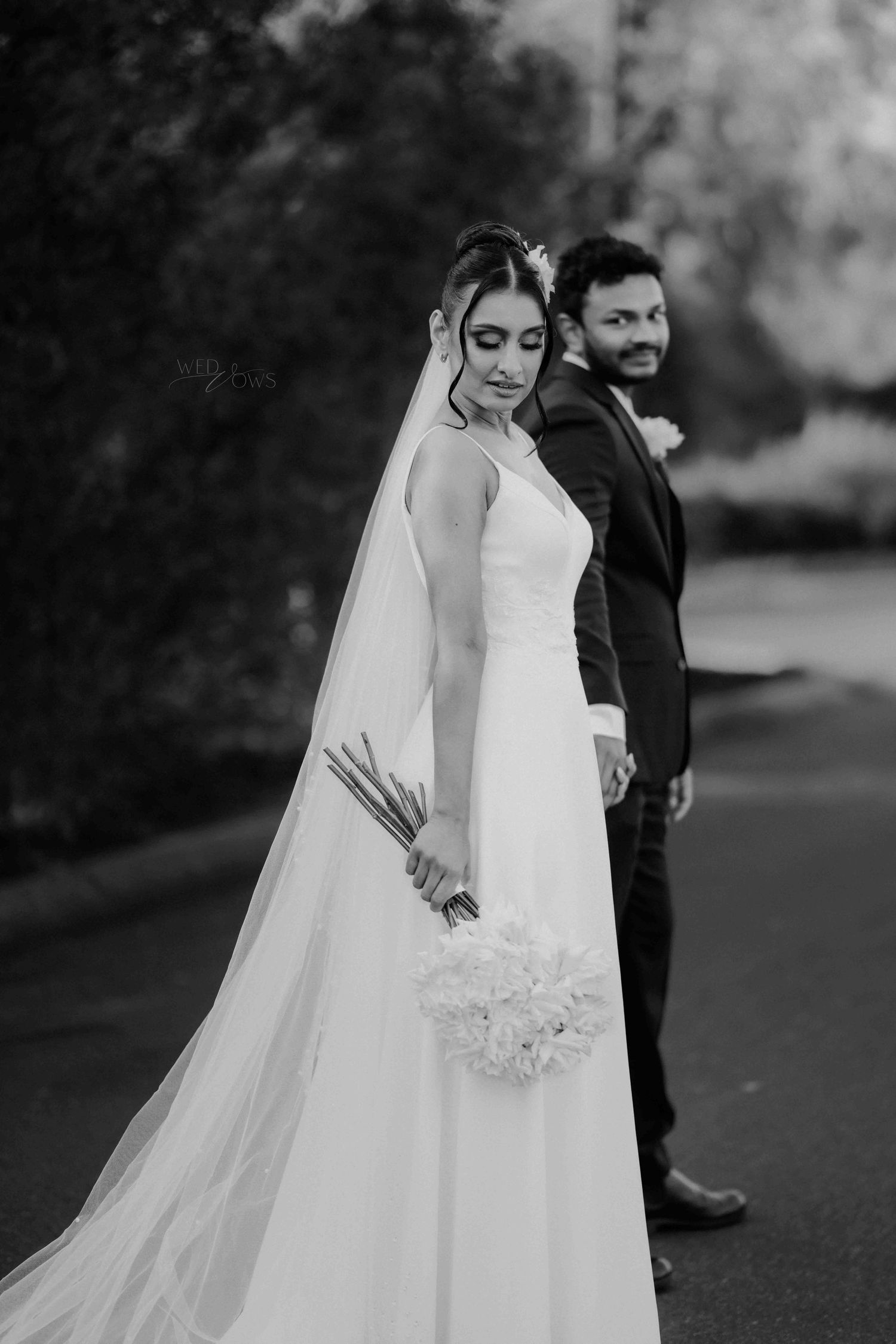 Black and white photo of bride in long veil holding bouquet, groom standing behind her outdoors.