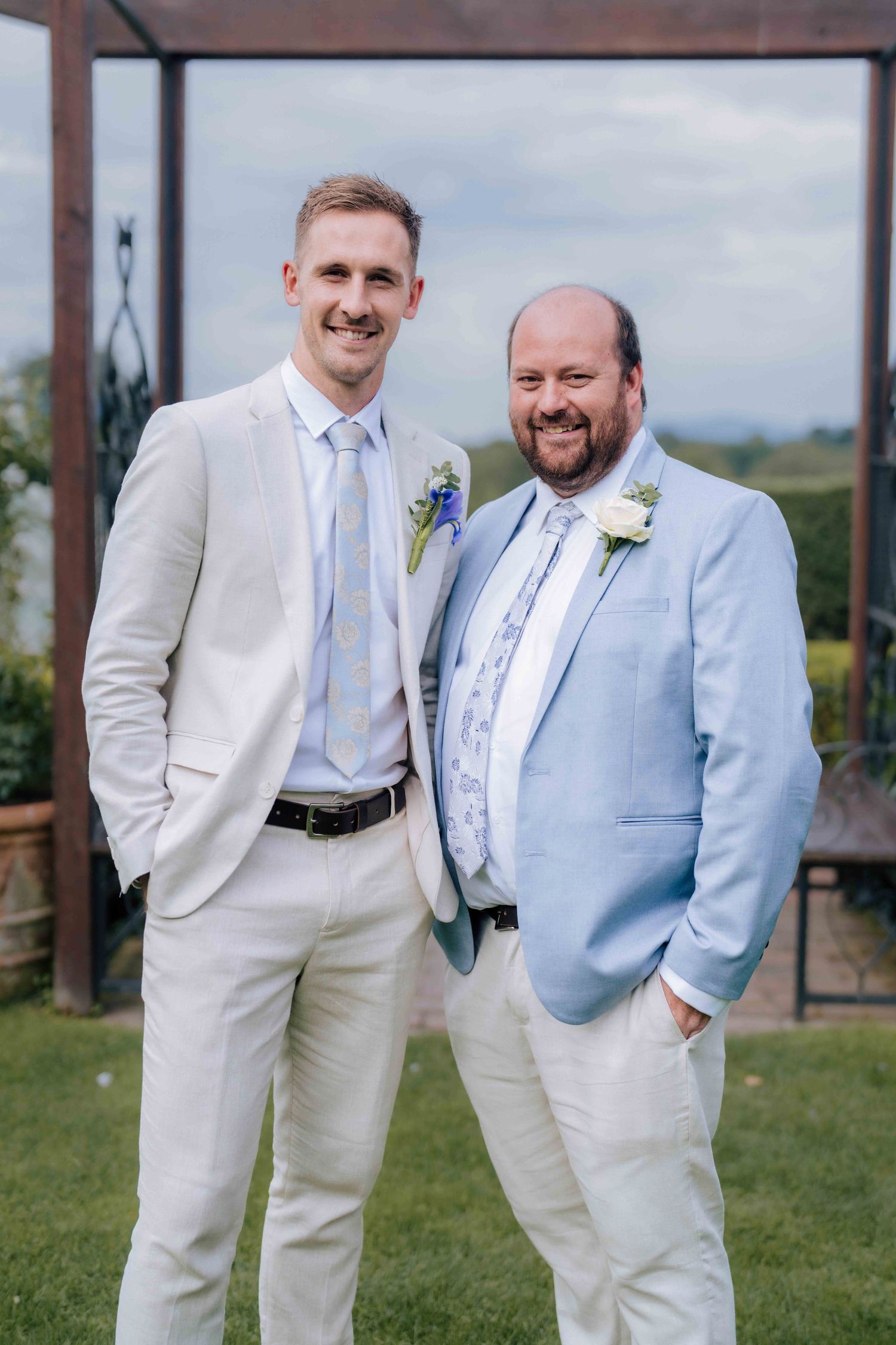 Tall groom in cream suit poses with older groomsman in blue blazer at outdoor wedding.