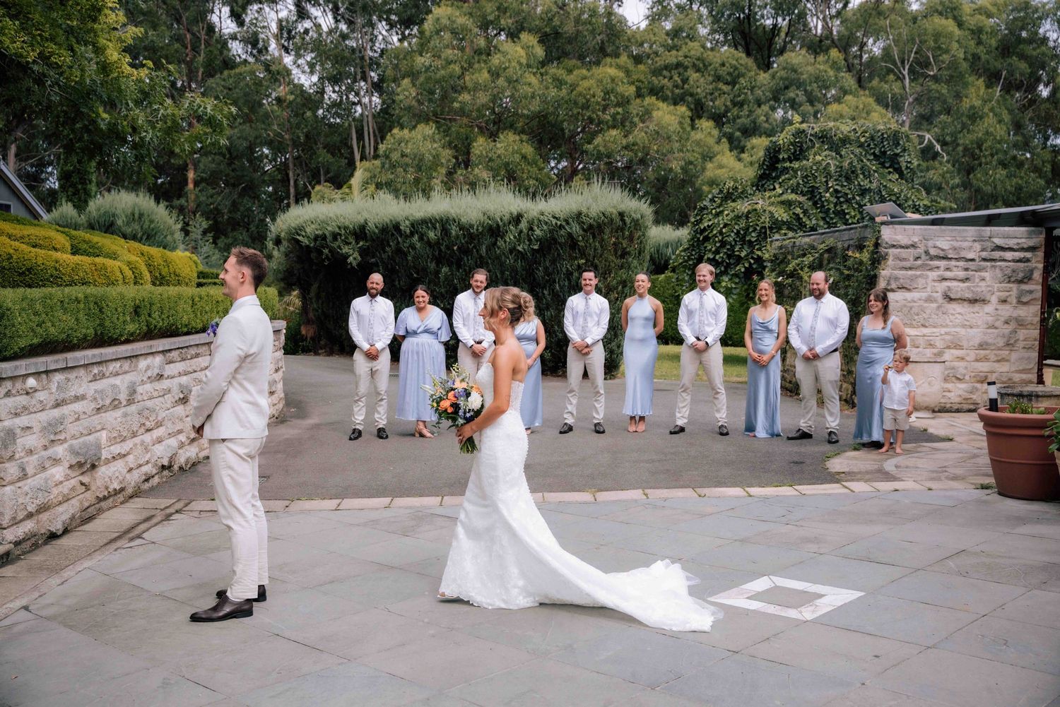 Bride in white gown approaches groom during outdoor first look with bridal party lined up behind.
