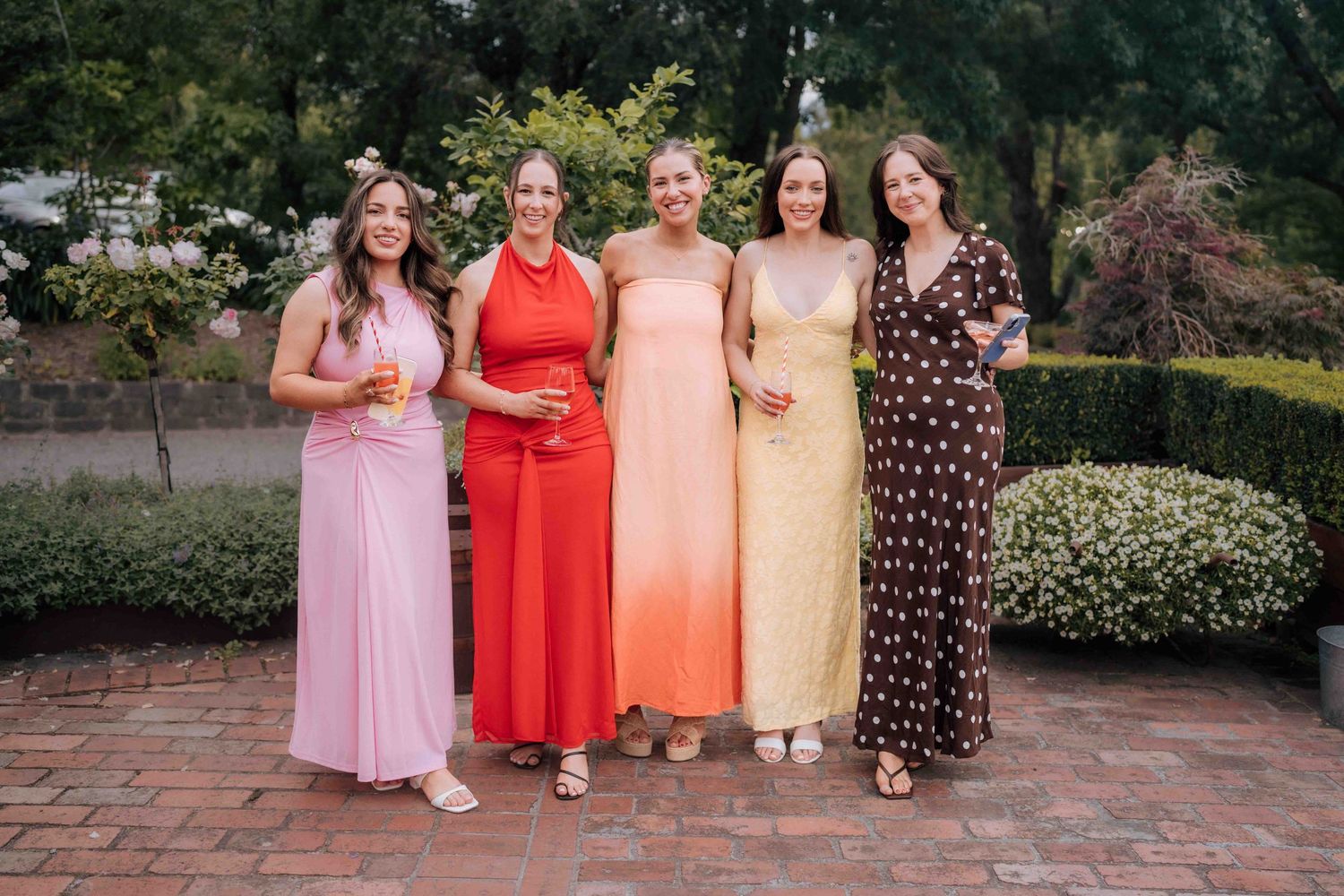 Five women in colorful formal dresses pose together at an outdoor wedding reception.
