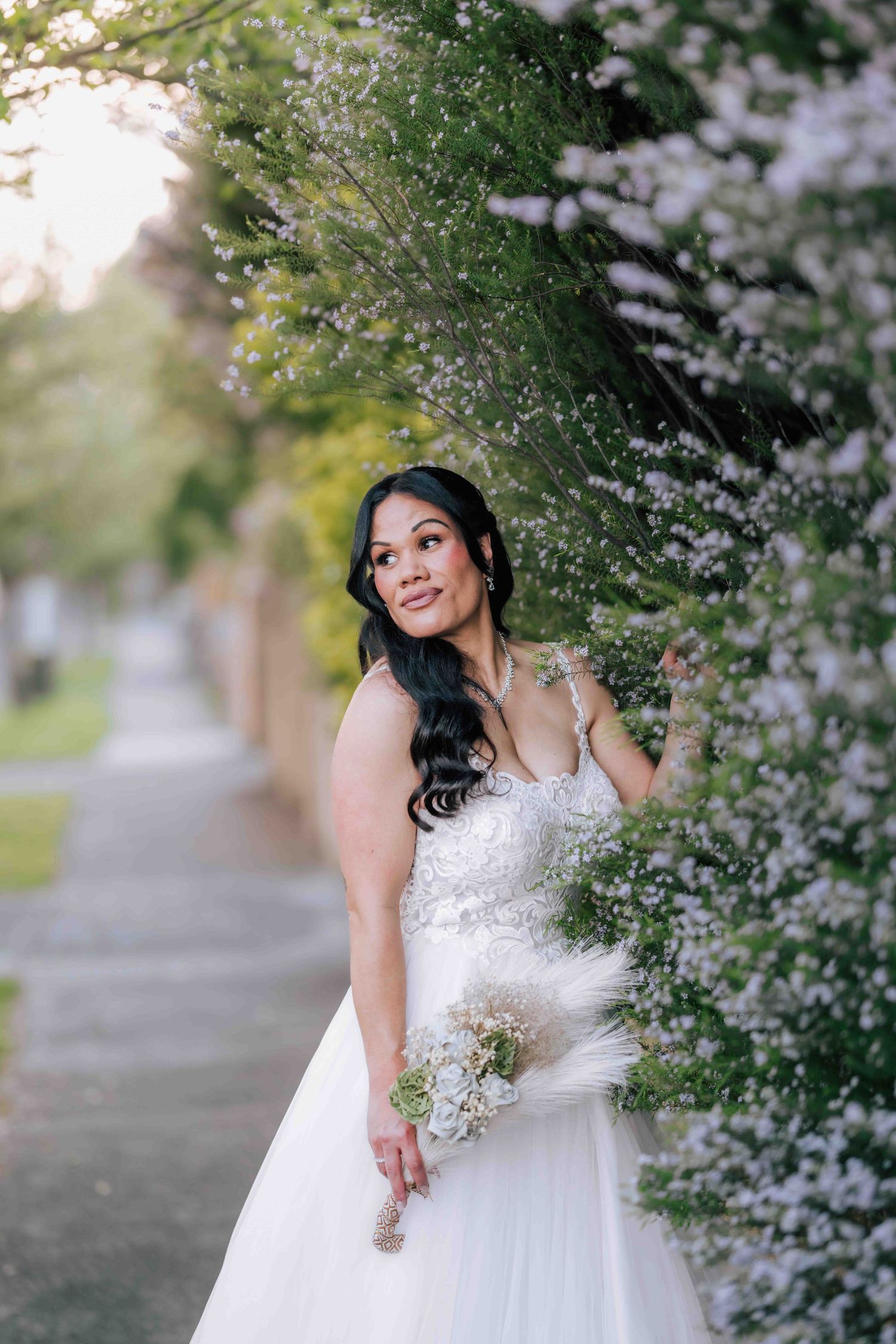 Radiant bride in a white ball gown posing among blooming white flowers on a garden path.