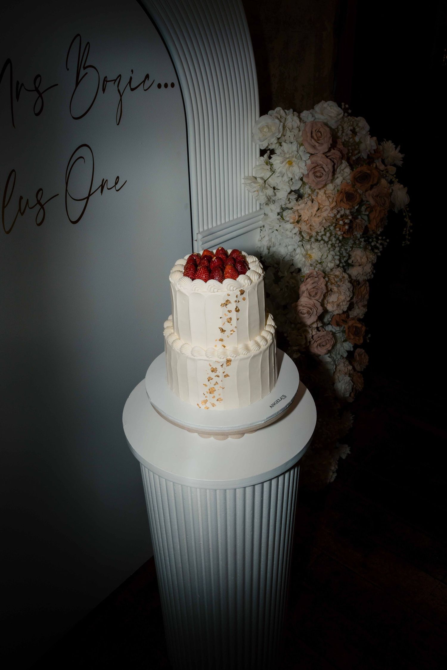 Two-tier white wedding cake topped with fresh strawberries on a white pedestal beside floral arrangement.