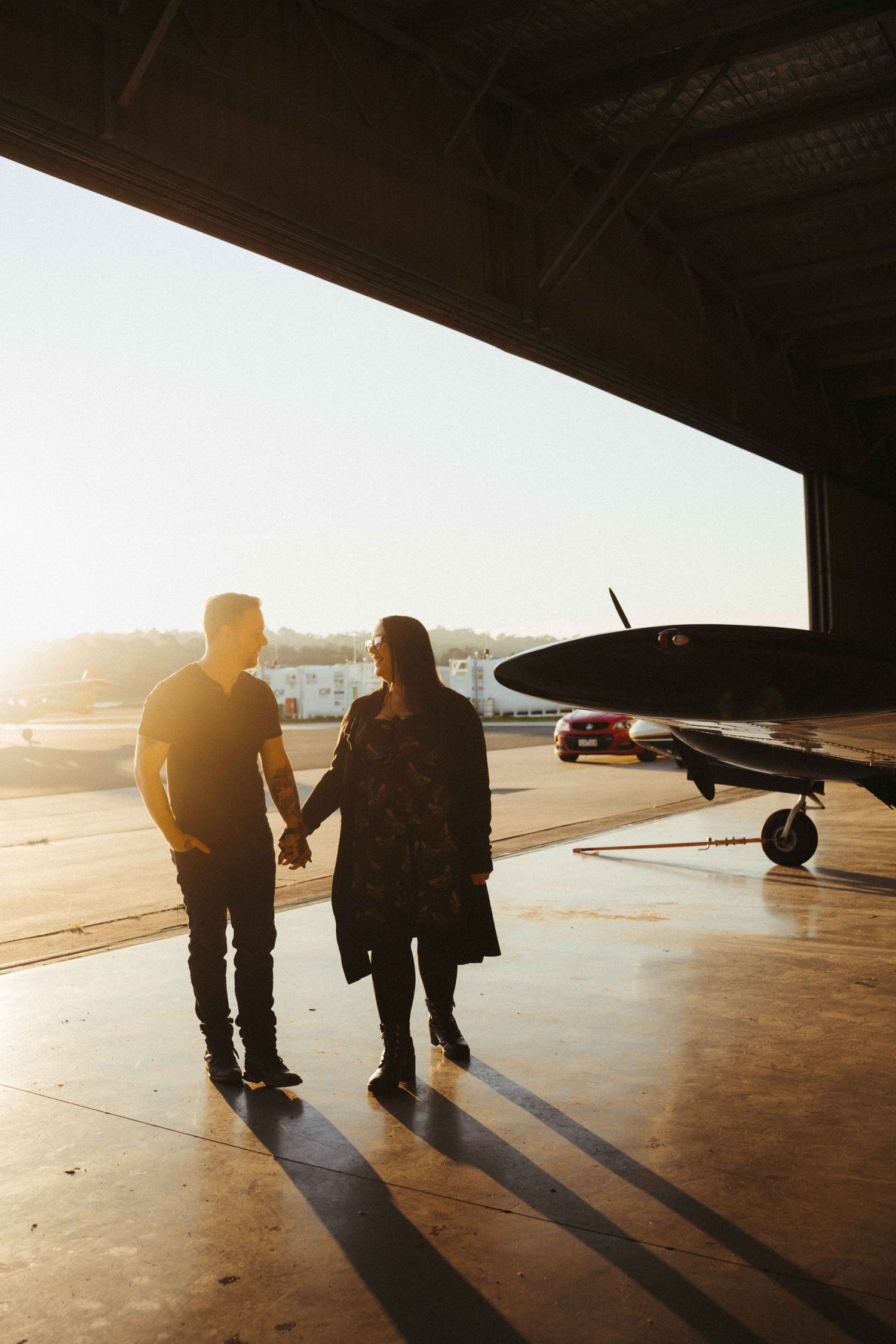 Couple holding hands walking through an airport hangar at golden hour with a small propeller plane nearby.