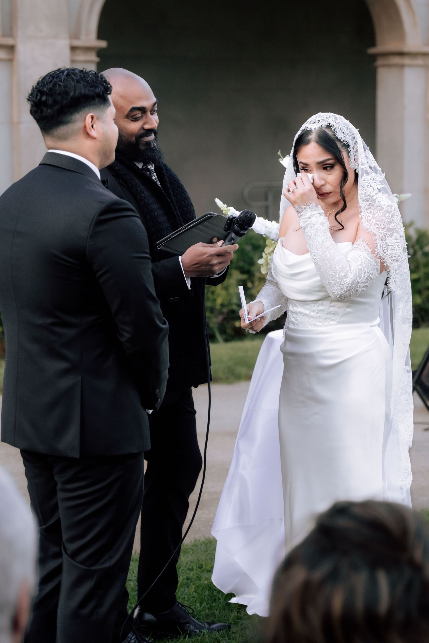 Emotional bride wiping tears while reading vows at elegant outdoor wedding ceremony.