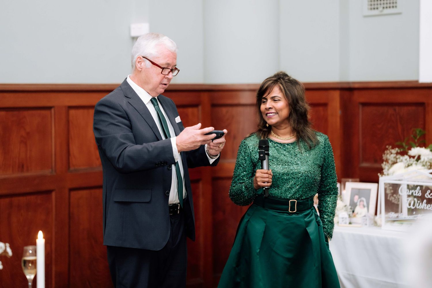 Man and woman in green dress laugh together during wedding reception speech at indoor venue.