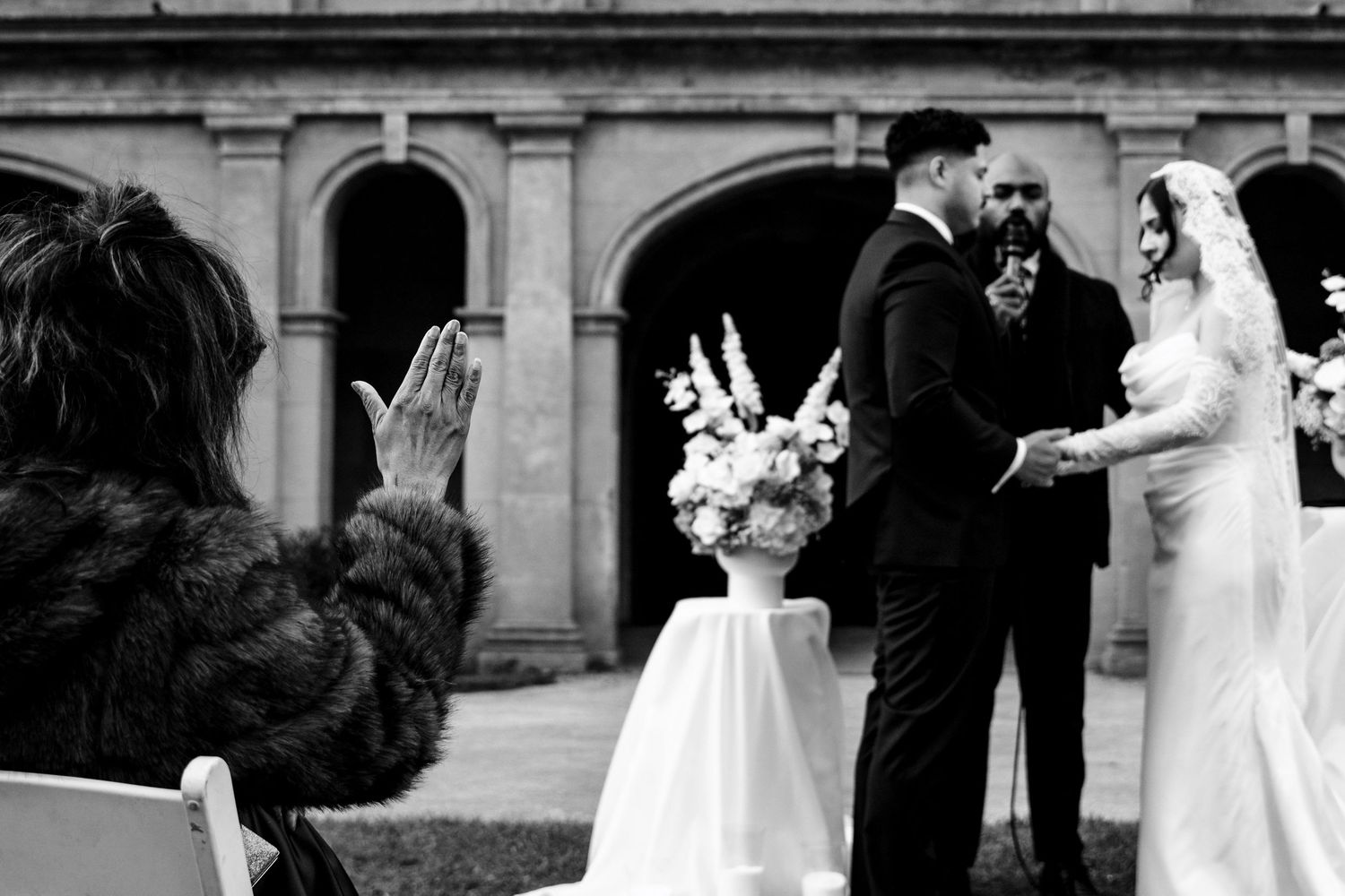 Black and white photo of a couple exchanging vows at an outdoor wedding ceremony with floral arrangements.