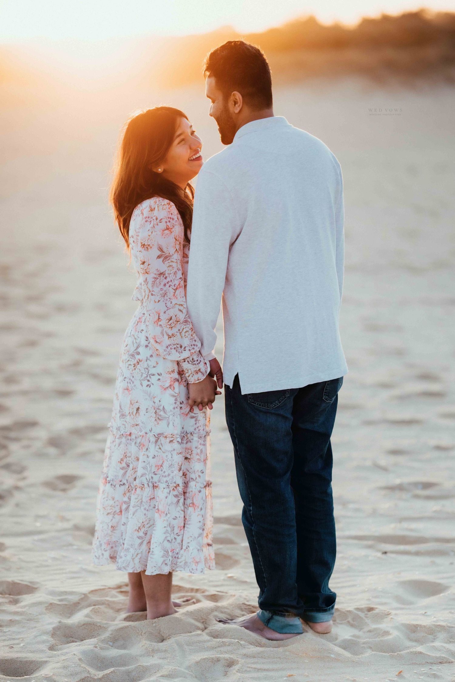Romantic couple holding hands on sandy beach at golden sunset, woman in floral dress gazing up at man in white shirt.
