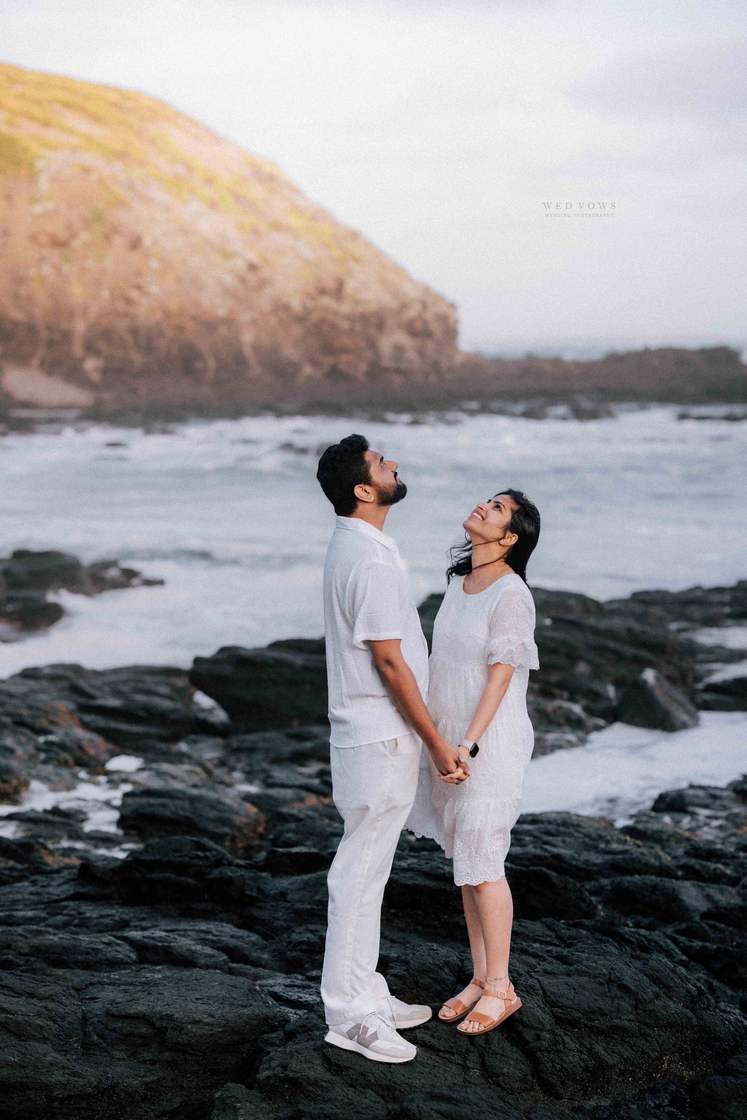 Couple in white holding hands and gazing at each other on rocky ocean shoreline.
