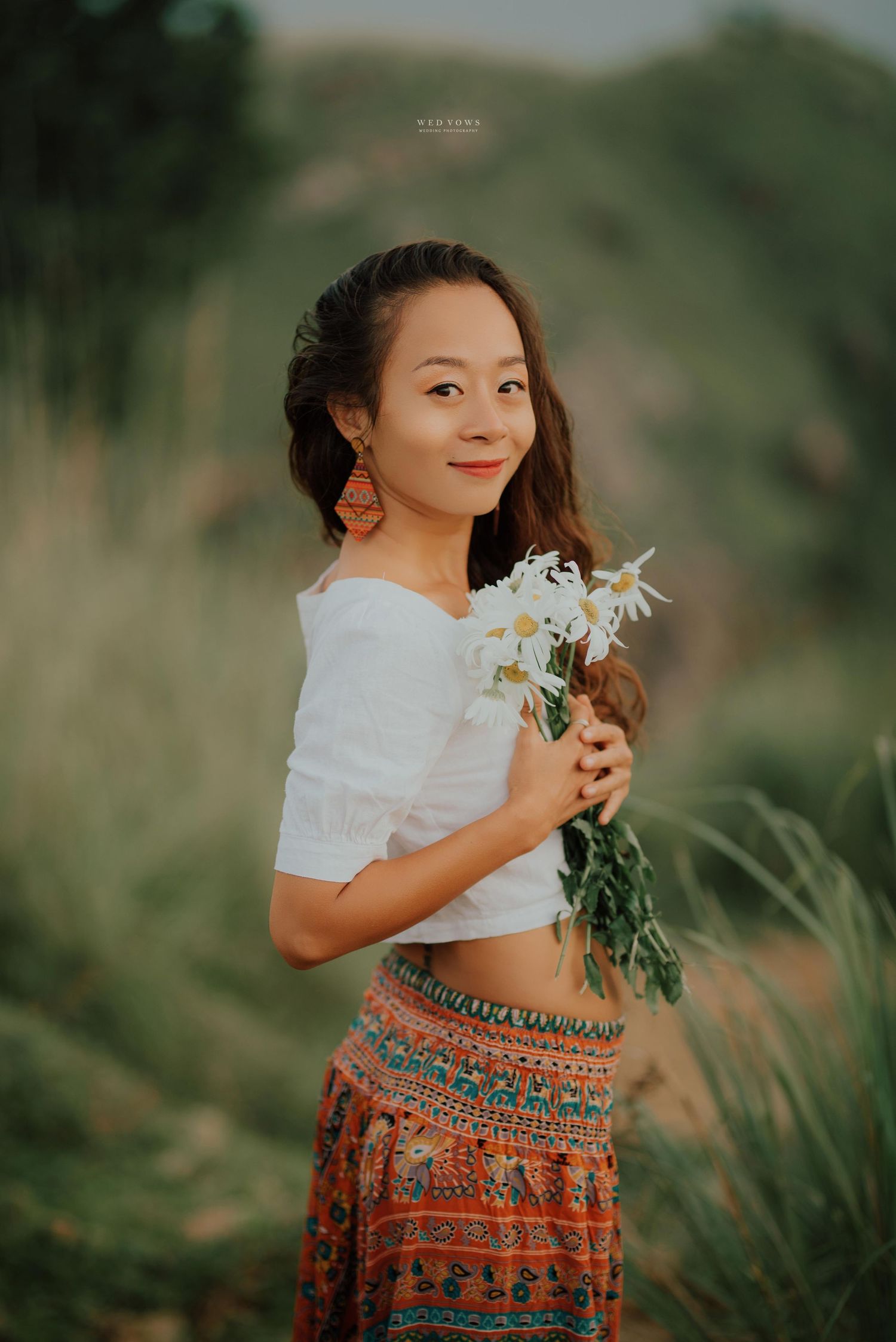 Smiling young woman holding white daisies in bohemian outfit among tall grasses.