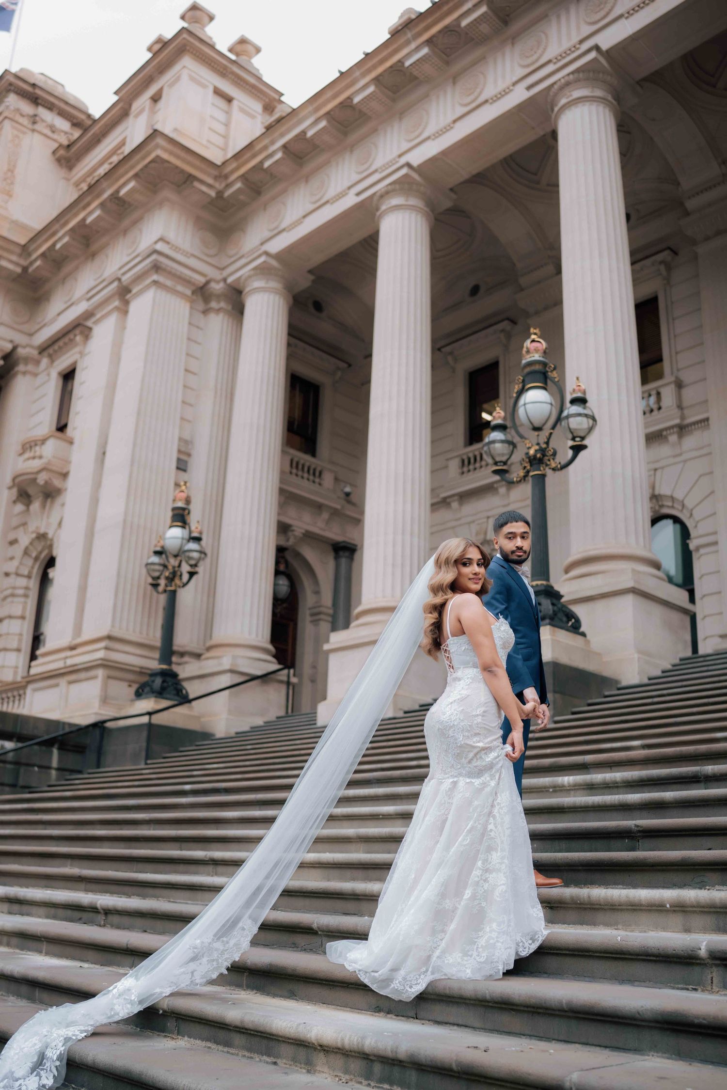 Bride in mermaid gown with long veil and groom in navy suit on grand building staircase.