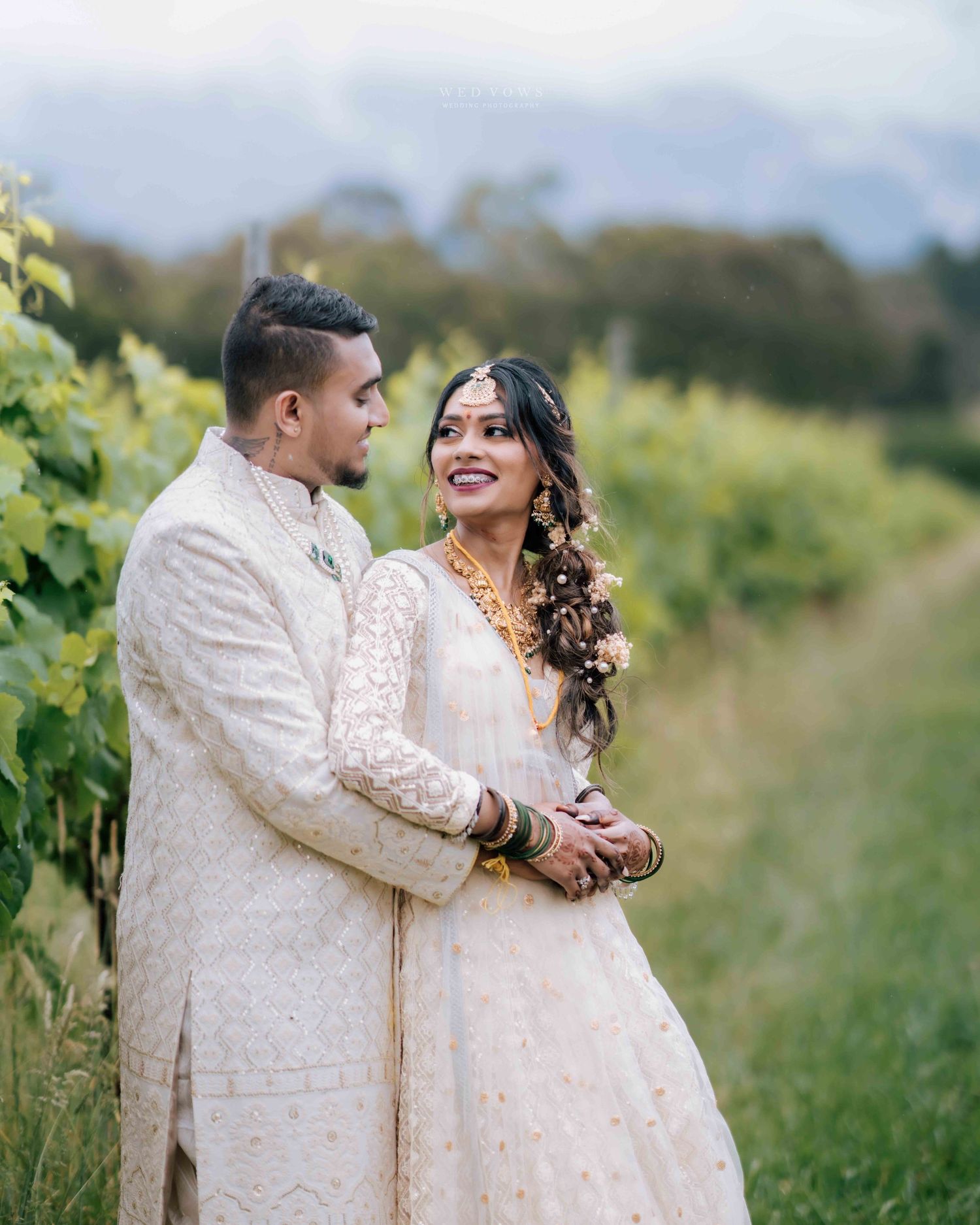 Indian wedding couple in matching white embroidered outfits sharing romantic moment in vineyard.