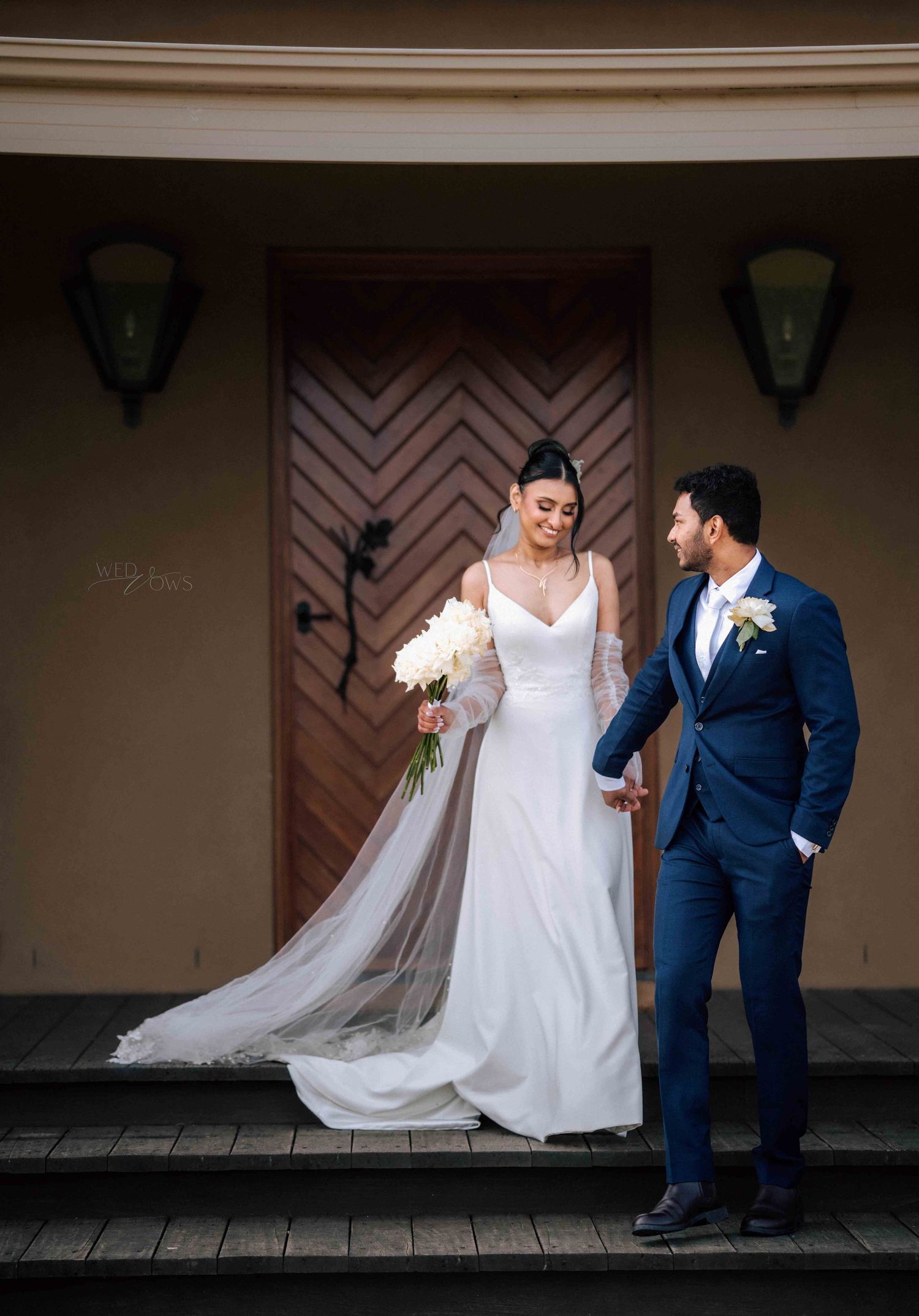 Bride and groom holding hands on steps before a wooden chevron door, bride in flowing white gown.