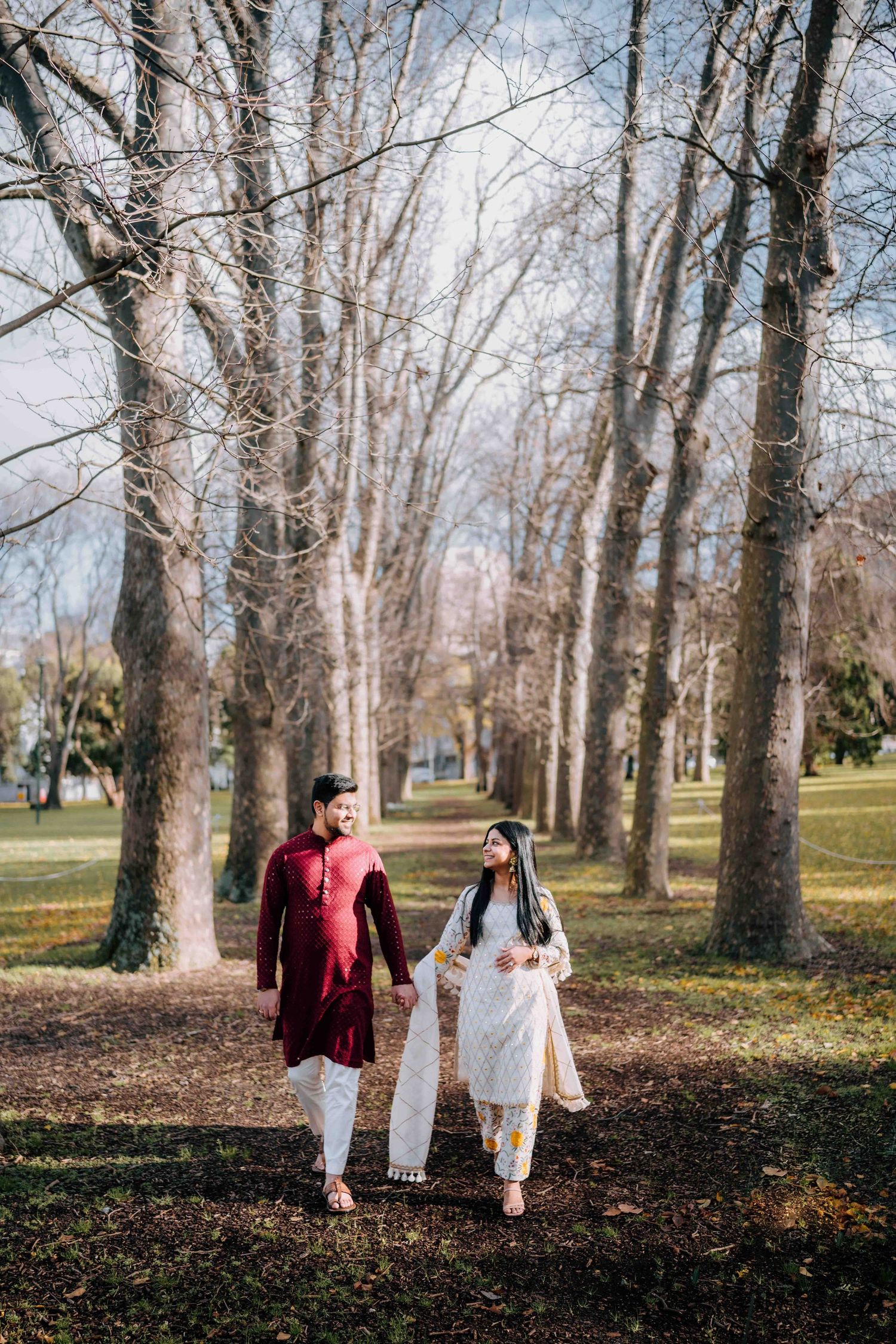 South Asian couple holding hands walking through tree-lined park path in traditional kurta outfits.