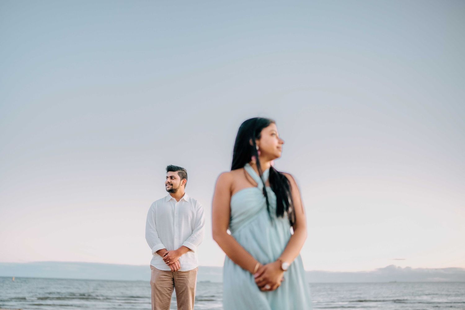Couple standing apart on a serene beach at dusk, woman in light blue dress, man in white shirt.