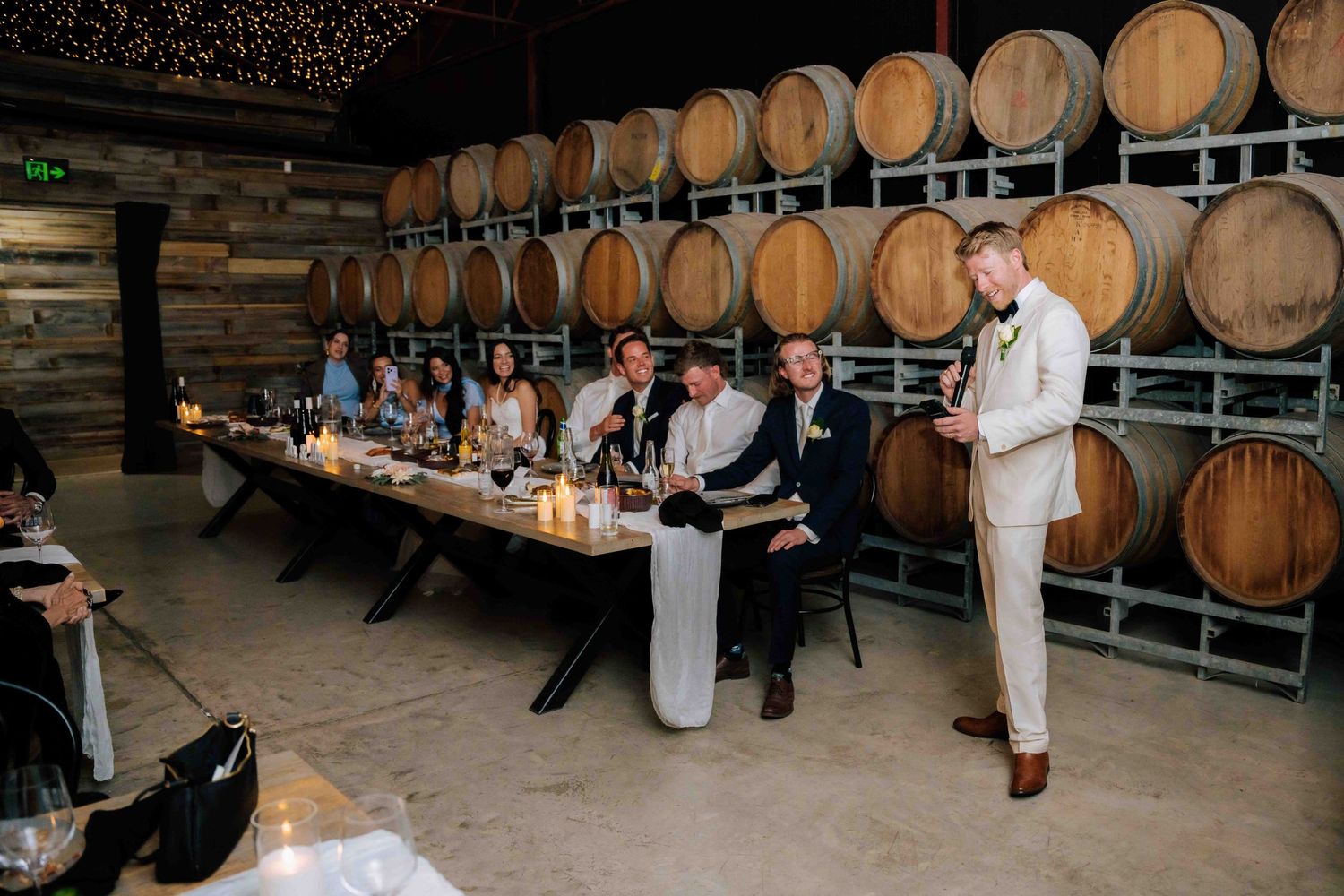Groom giving speech in winery barrel room during wedding reception dinner with guests seated.