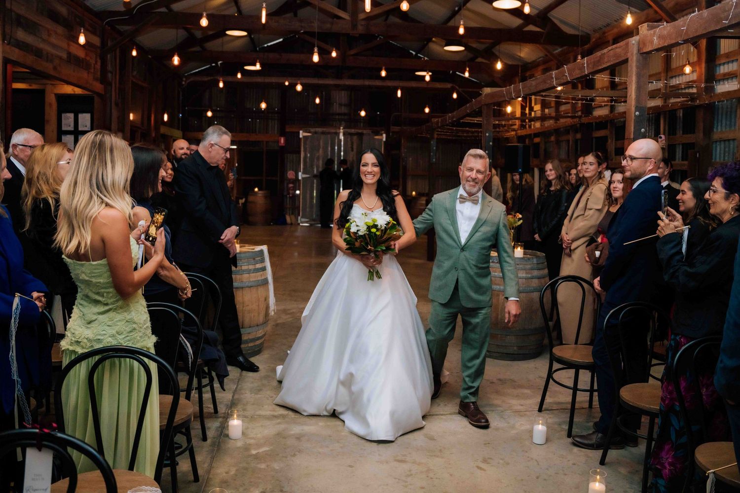 Bride and groom walk joyfully down the aisle in a rustic barn wedding venue surrounded by cheering guests.