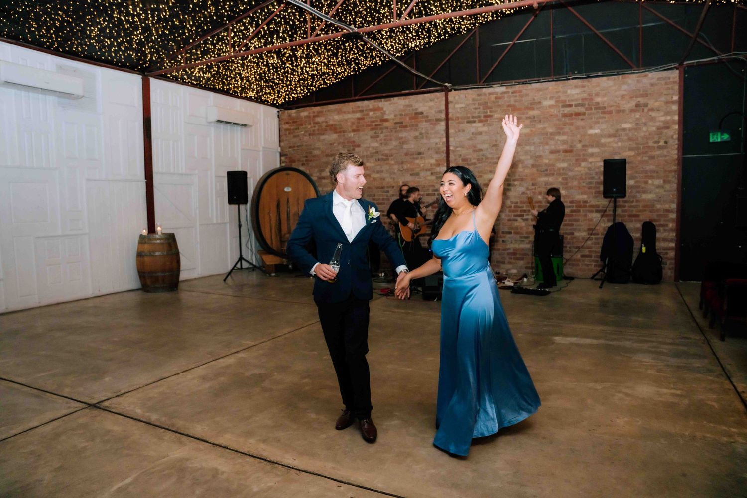 Couple dancing at wedding reception in rustic venue with string lights.