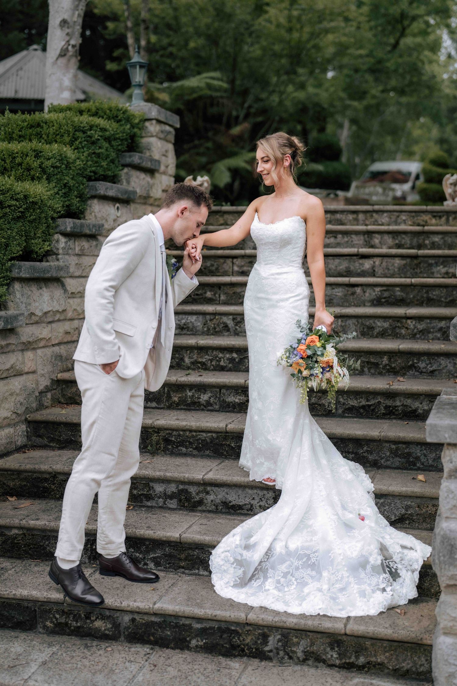 Groom in white suit kissing bride's hand on stone steps as she smiles holding colorful bouquet.