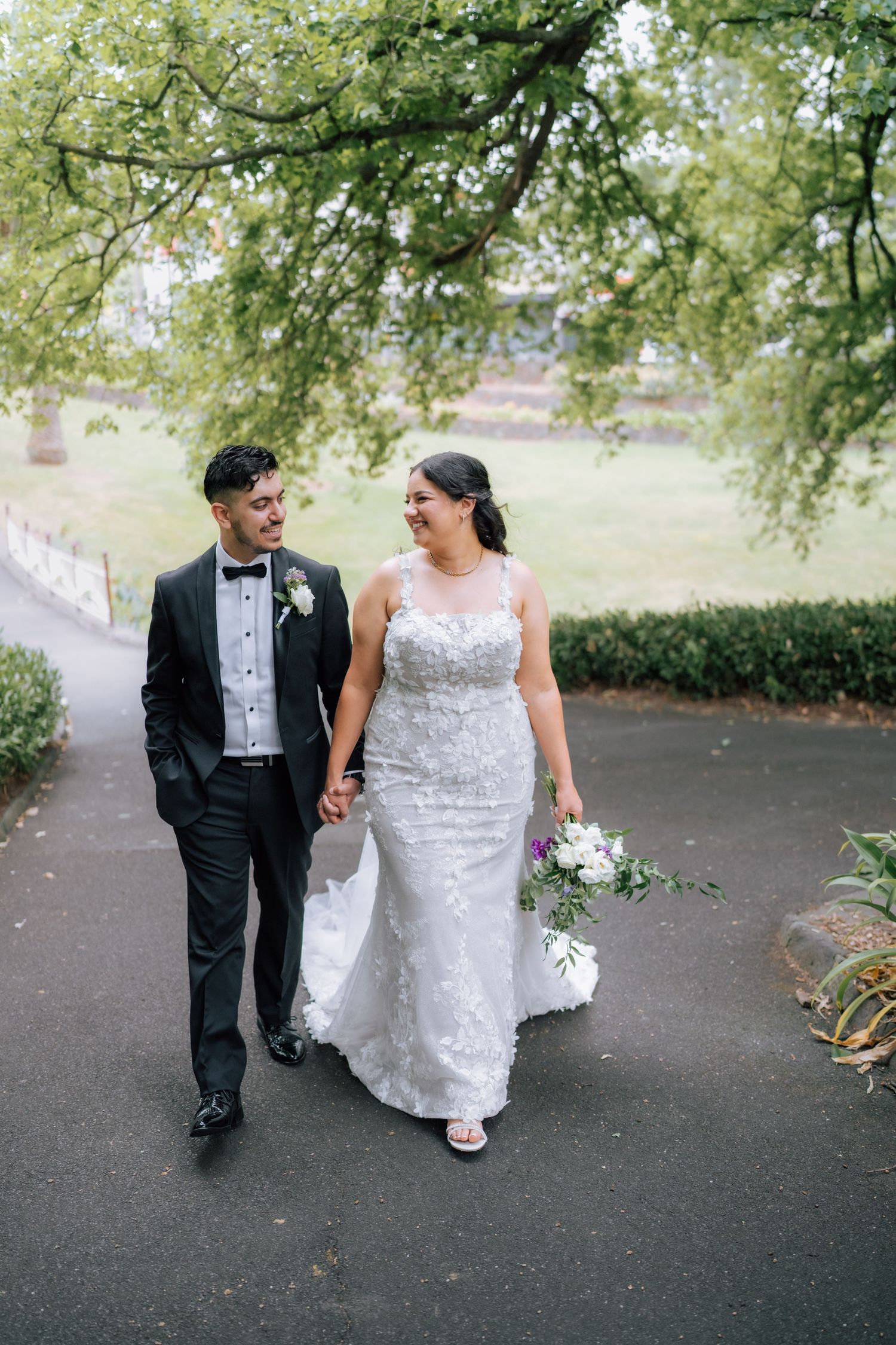 Bride in lace gown and groom in black tuxedo walking hand in hand through park.