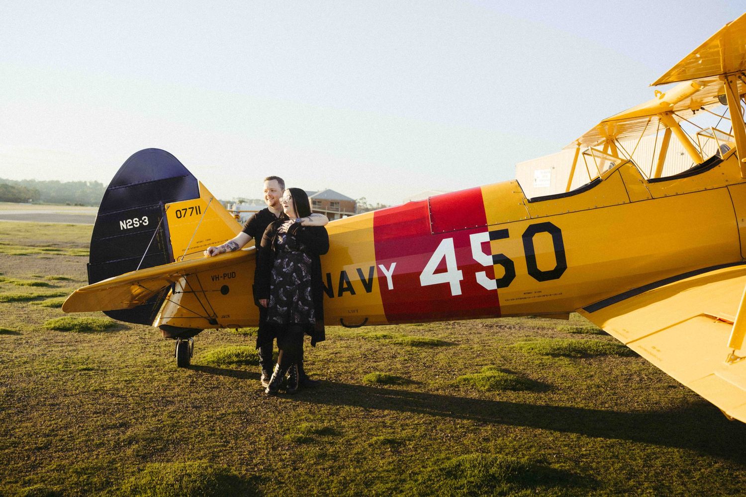 Couple posing beside a vintage yellow Navy 450 biplane on a grass airfield at sunset.