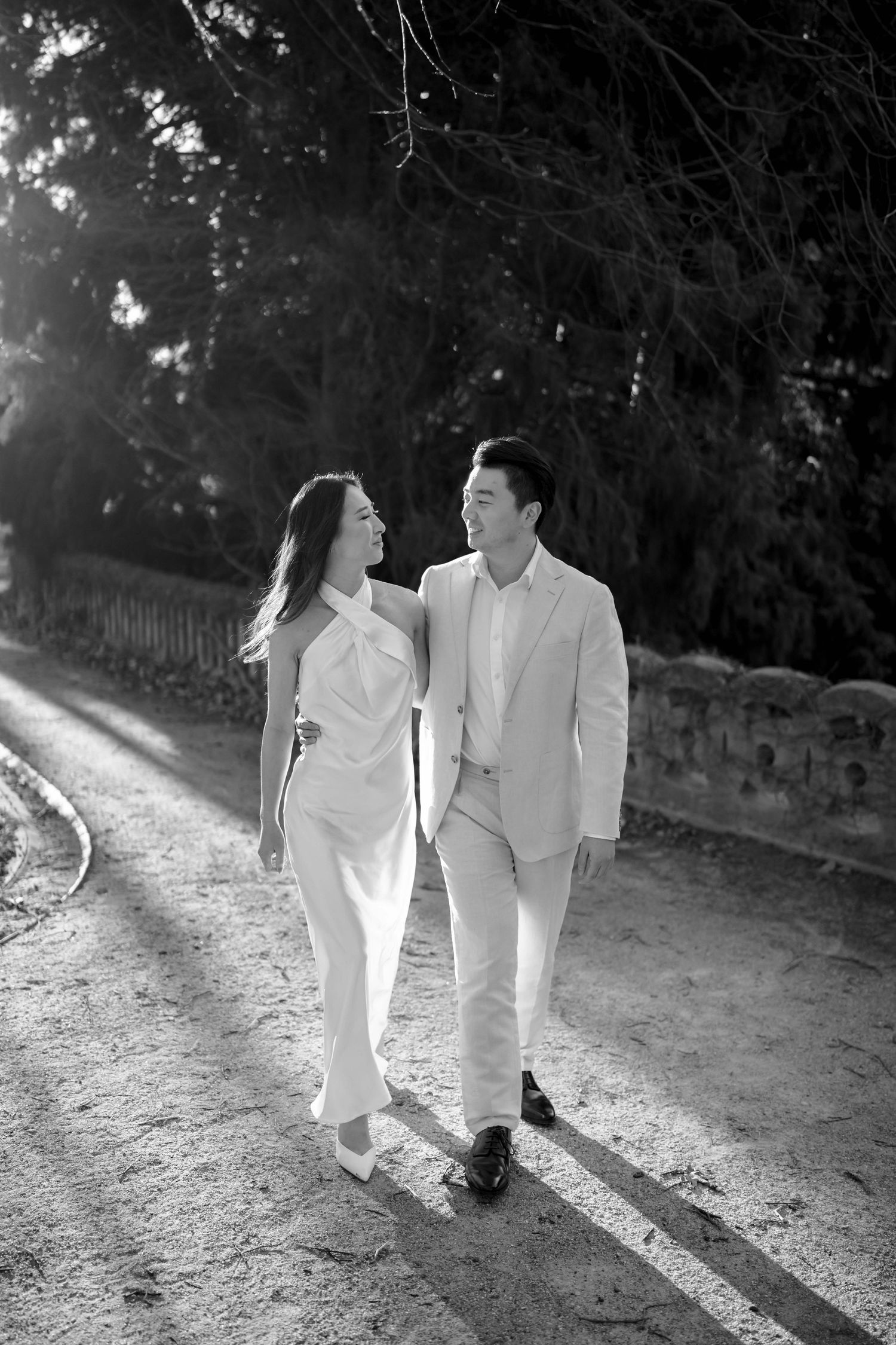 Black and white photo of a couple in white outfits walking and smiling at each other on a sunlit path.
