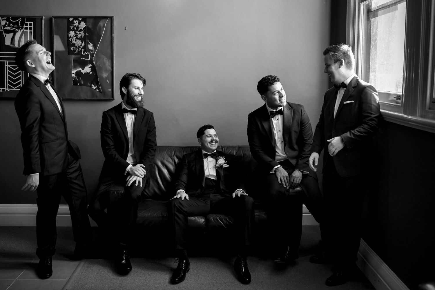 Black and white photo of five groomsmen in tuxedos laughing together in a stylish room.