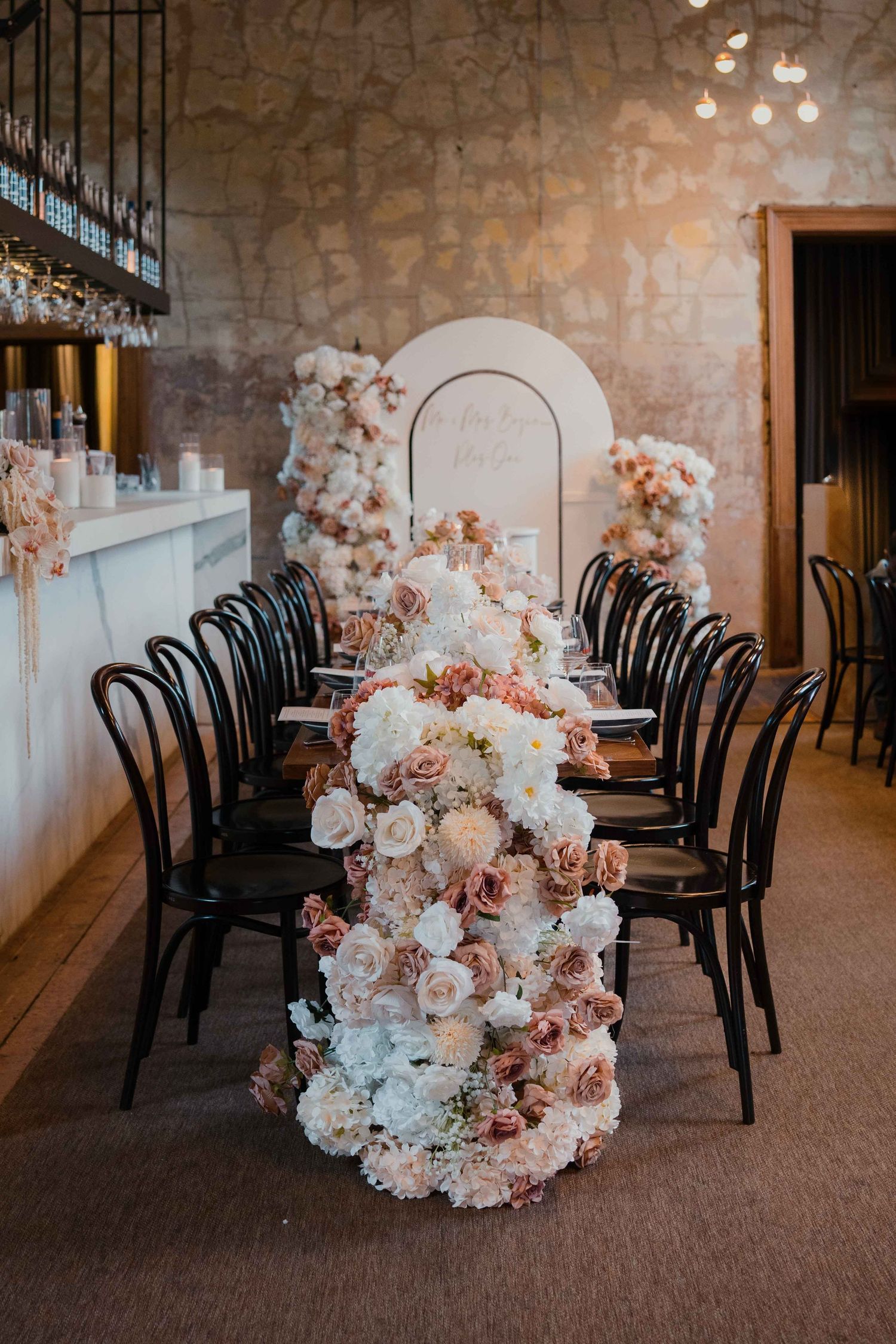 Stunning wedding reception table with overflowing blush and white rose floral runner, black chairs, and arched backdrop in stone venue.