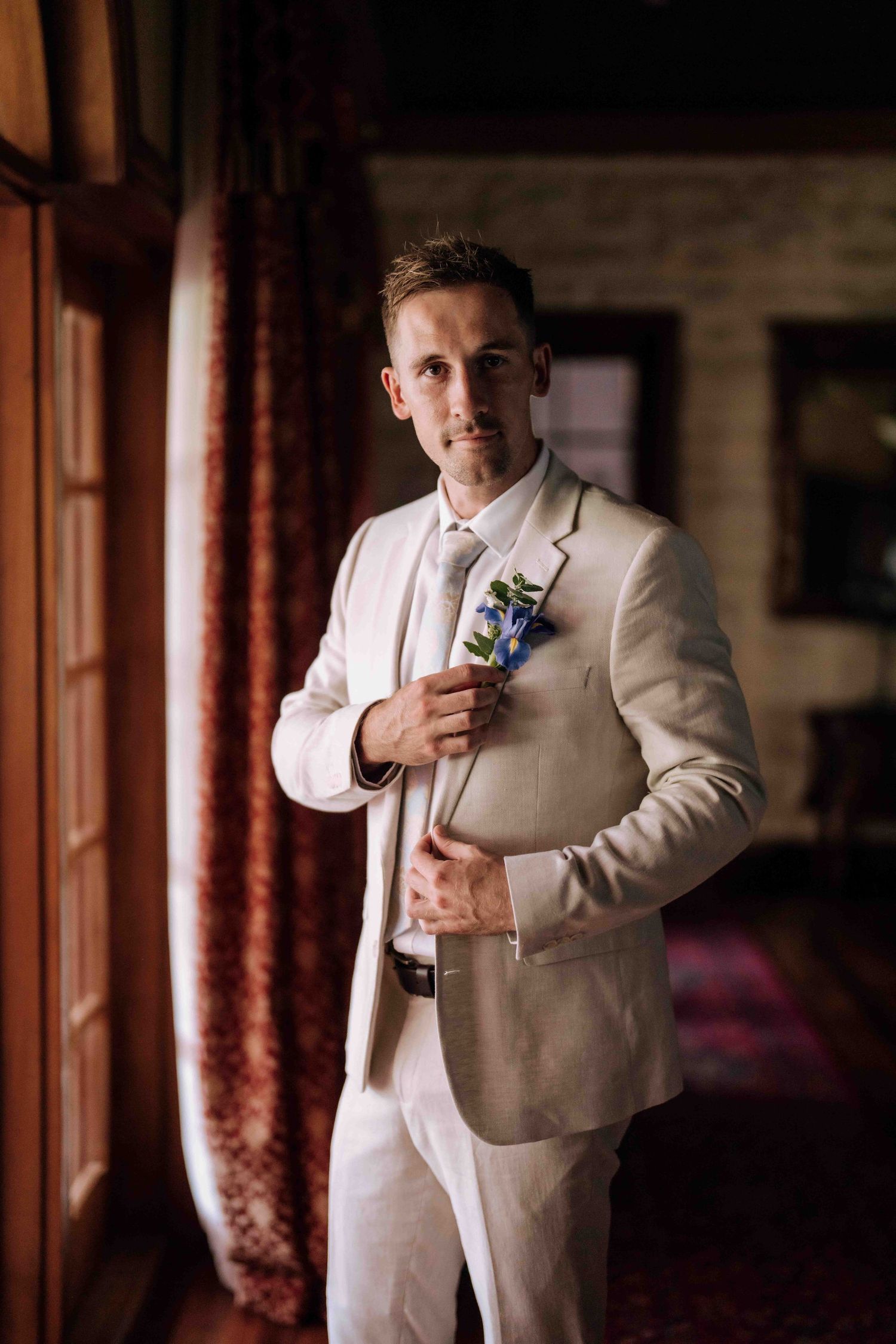 Dapper groom in cream suit adjusting blue boutonniere while posing by window on wedding day.