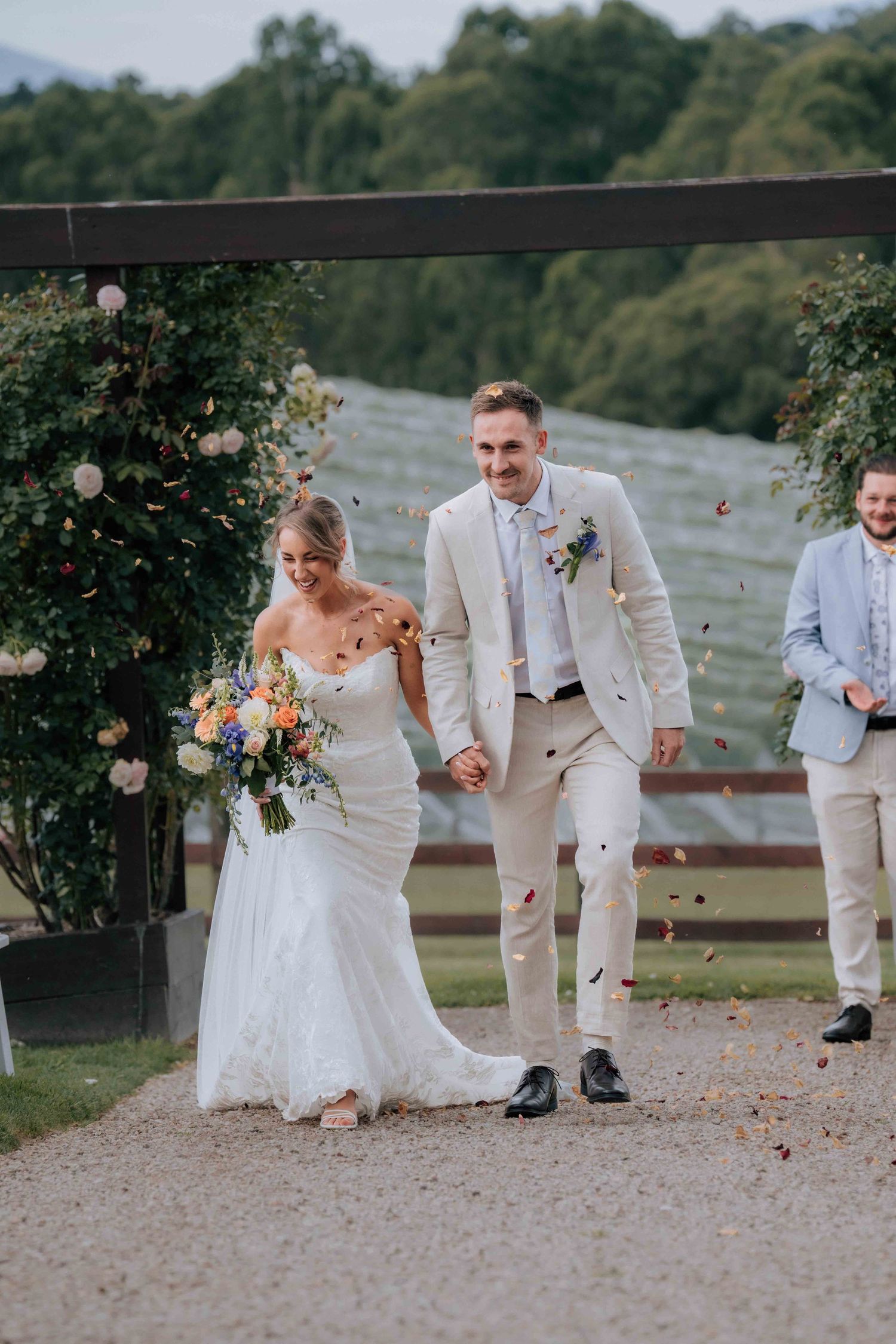Happy bride and groom exit wedding ceremony through floral arch as guests throw colorful confetti petals.