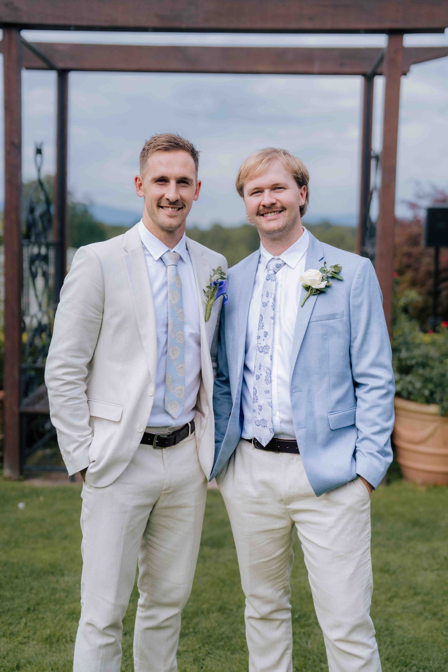 Groom and best man in cream and blue suits with boutonnieres posing outdoors at wedding venue.