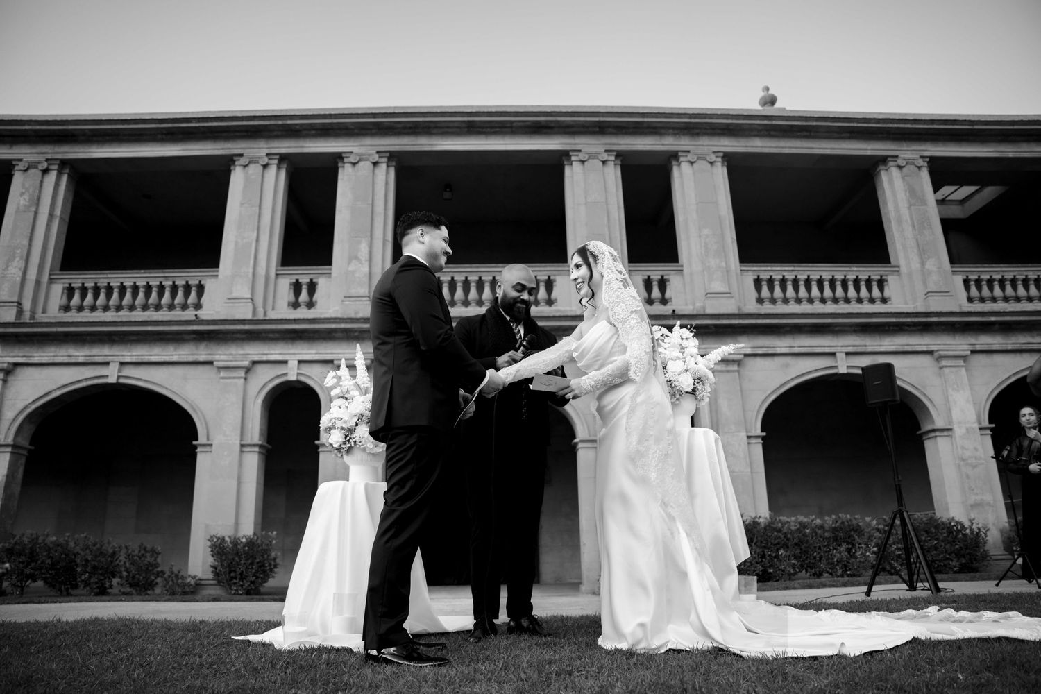 Black and white photo of bride and groom exchanging vows with officiant at elegant outdoor ceremony.